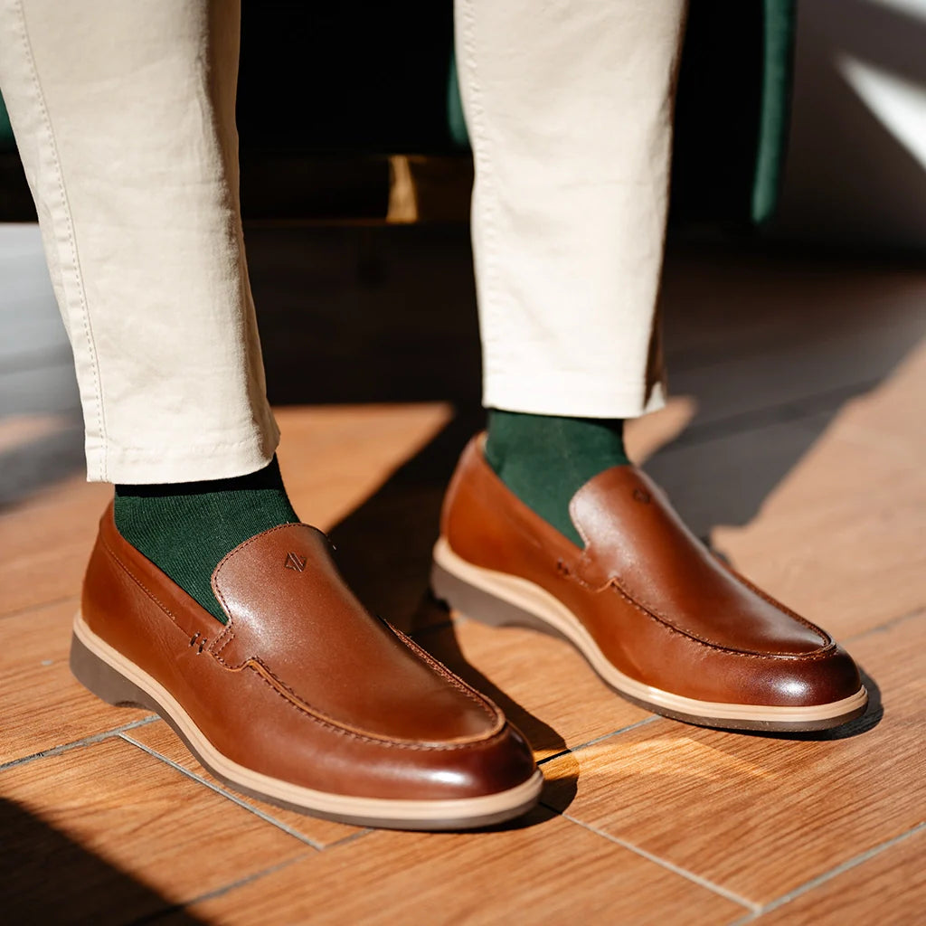 Brown loafers worn with white pants and green socks on a wooden floor.
