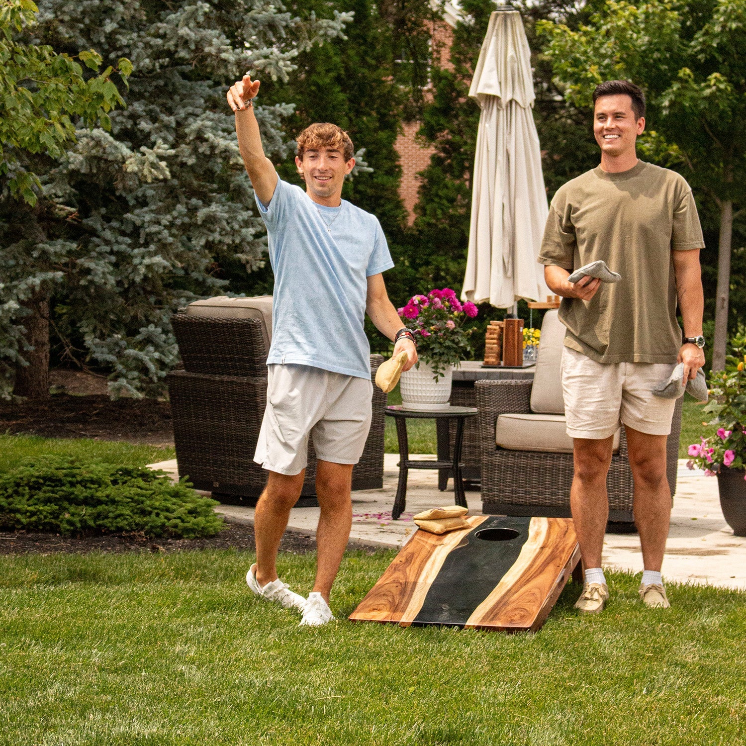 Two men playing cornhole in a backyard setting with outdoor furniture and plants.