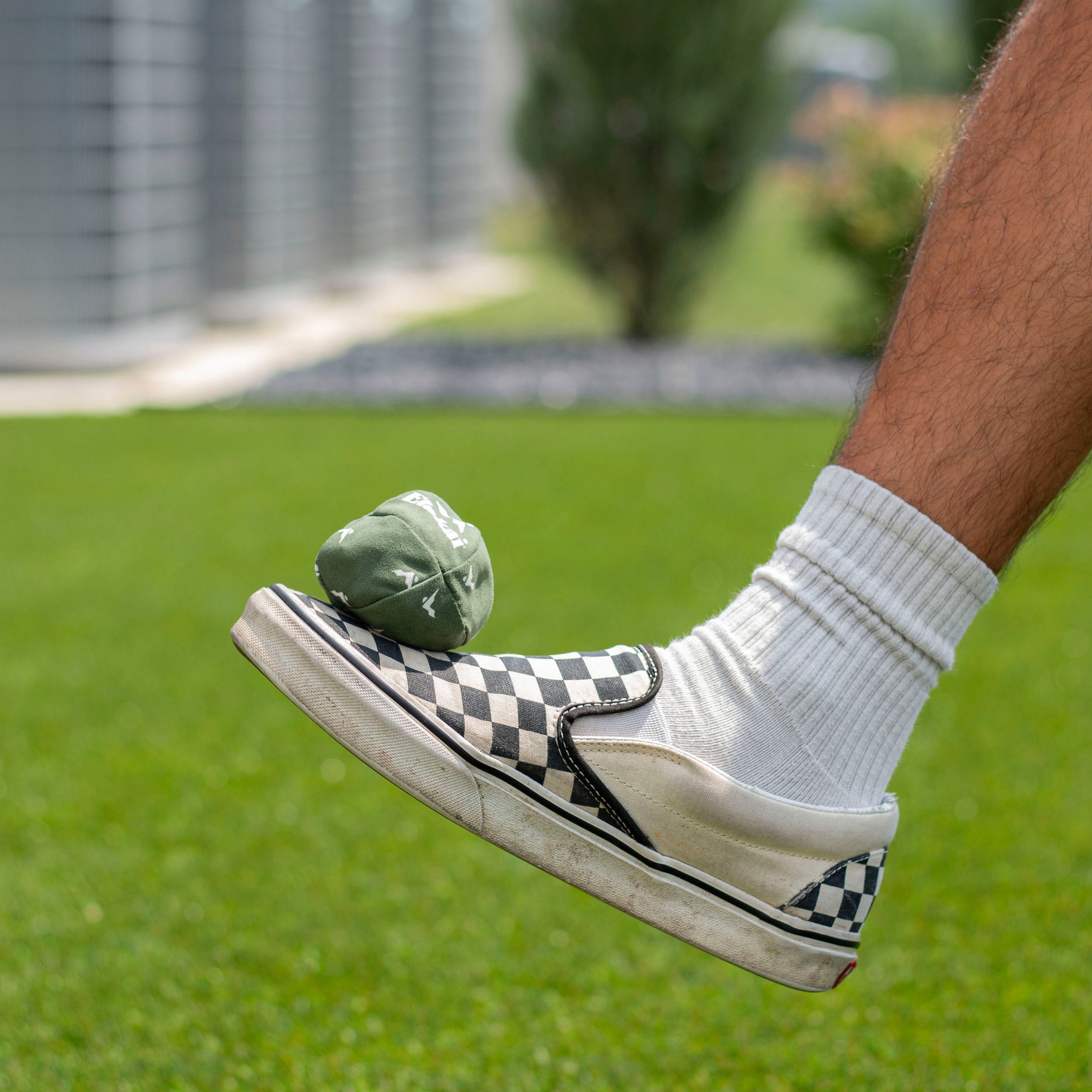 Person wearing a checkered sneaker with a white sock, stepping on a green bag on grass.