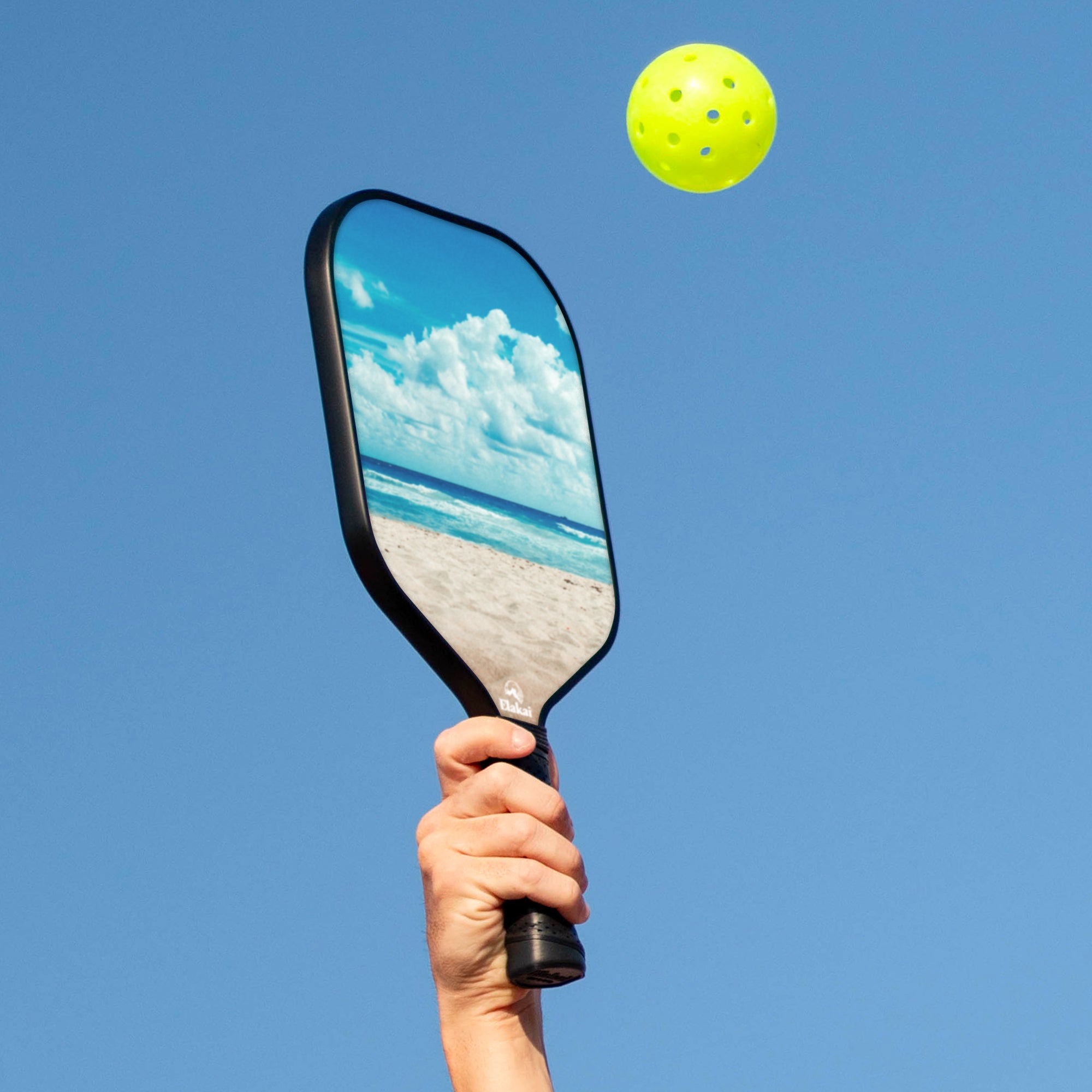 Hand holding a paddle with a beach reflection, next to a yellow ball against a clear blue sky.