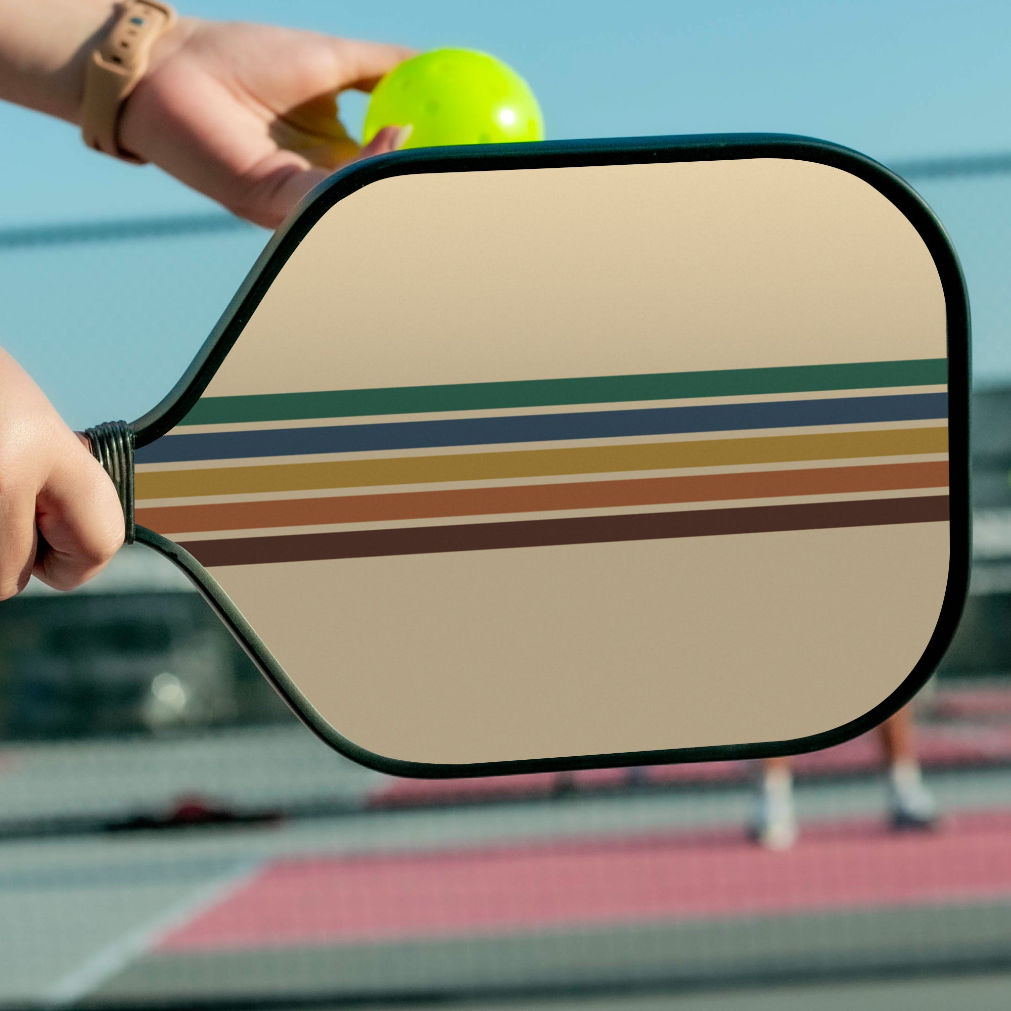 Close-up of a striped pickleball paddle with a blurred background of a pickleball court.