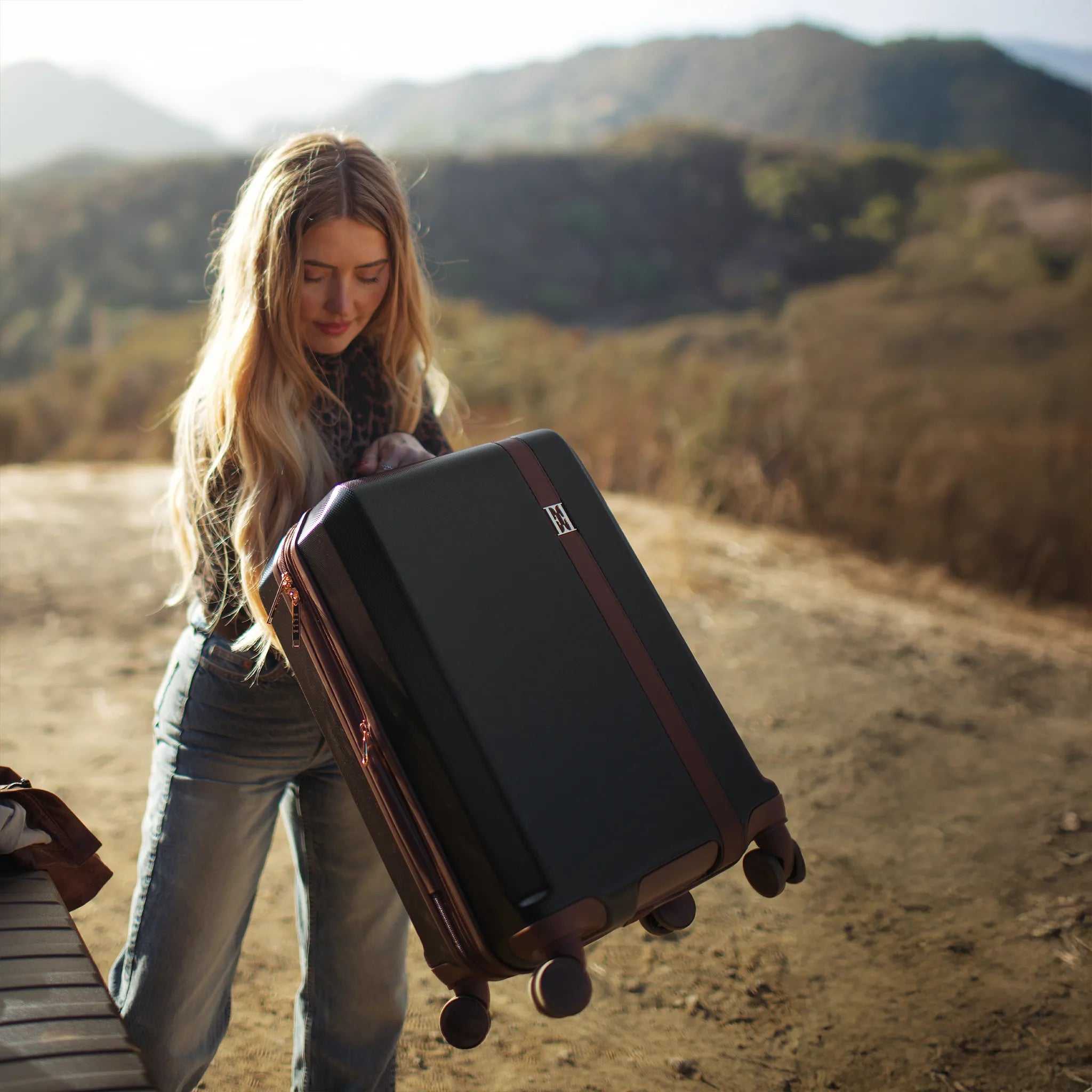 Woman holding a suitcase in a desert-like landscape with mountains in the background