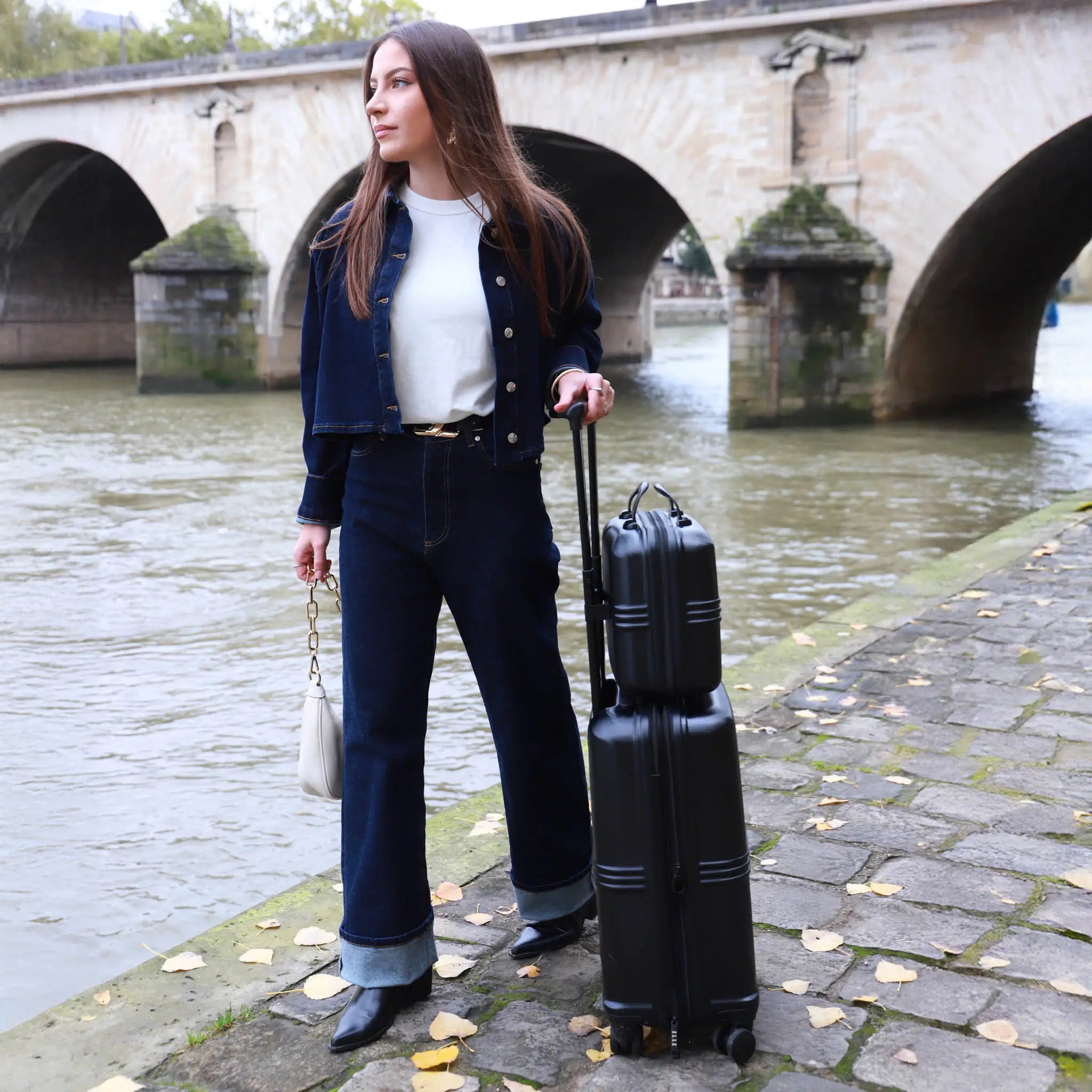 Woman with a suitcase standing by a river with a bridge in the background