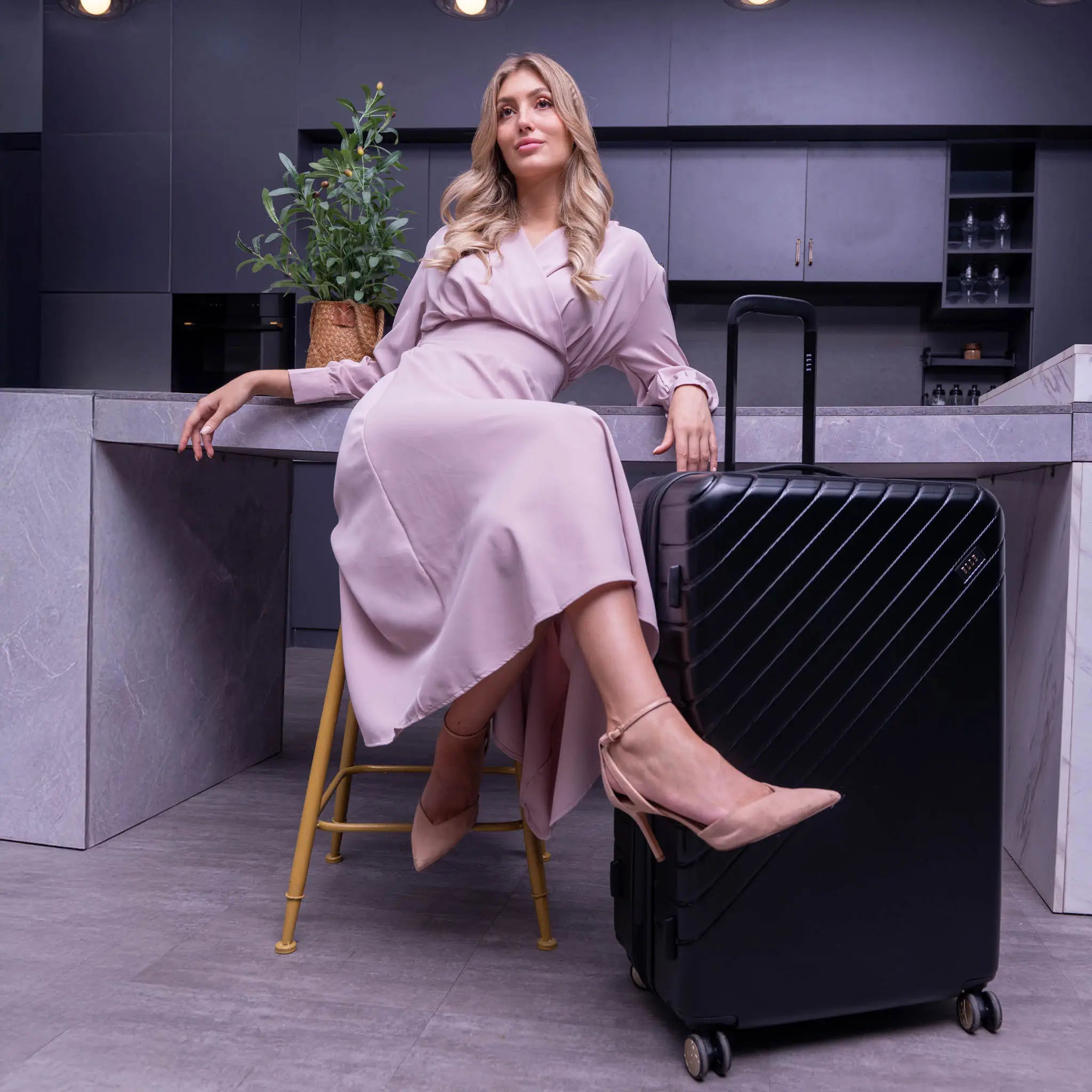 Woman in a pink dress sitting next to a black suitcase in a modern kitchen.