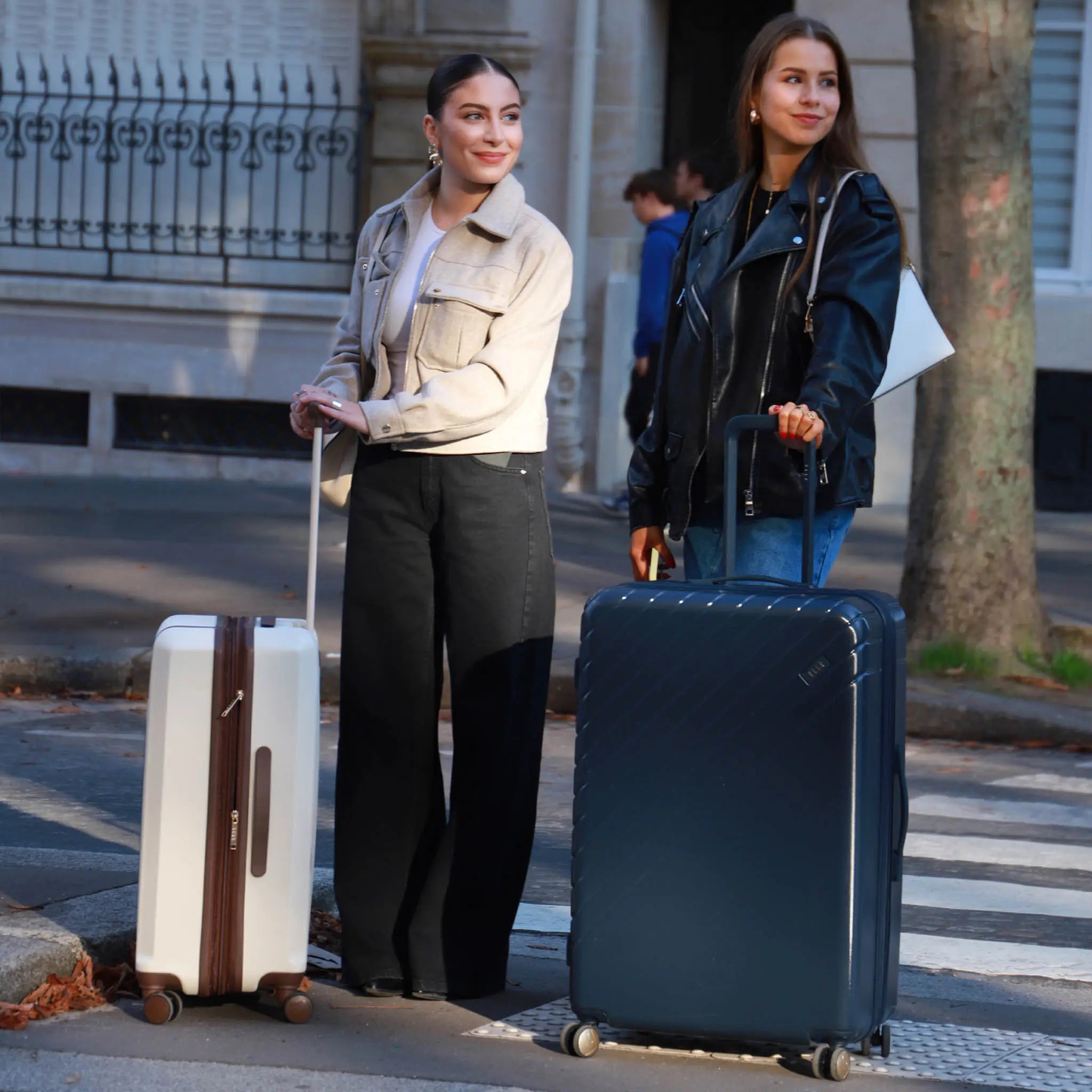 Two women with suitcases standing on a street, one holding a white suitcase and the other a blue suitcase.