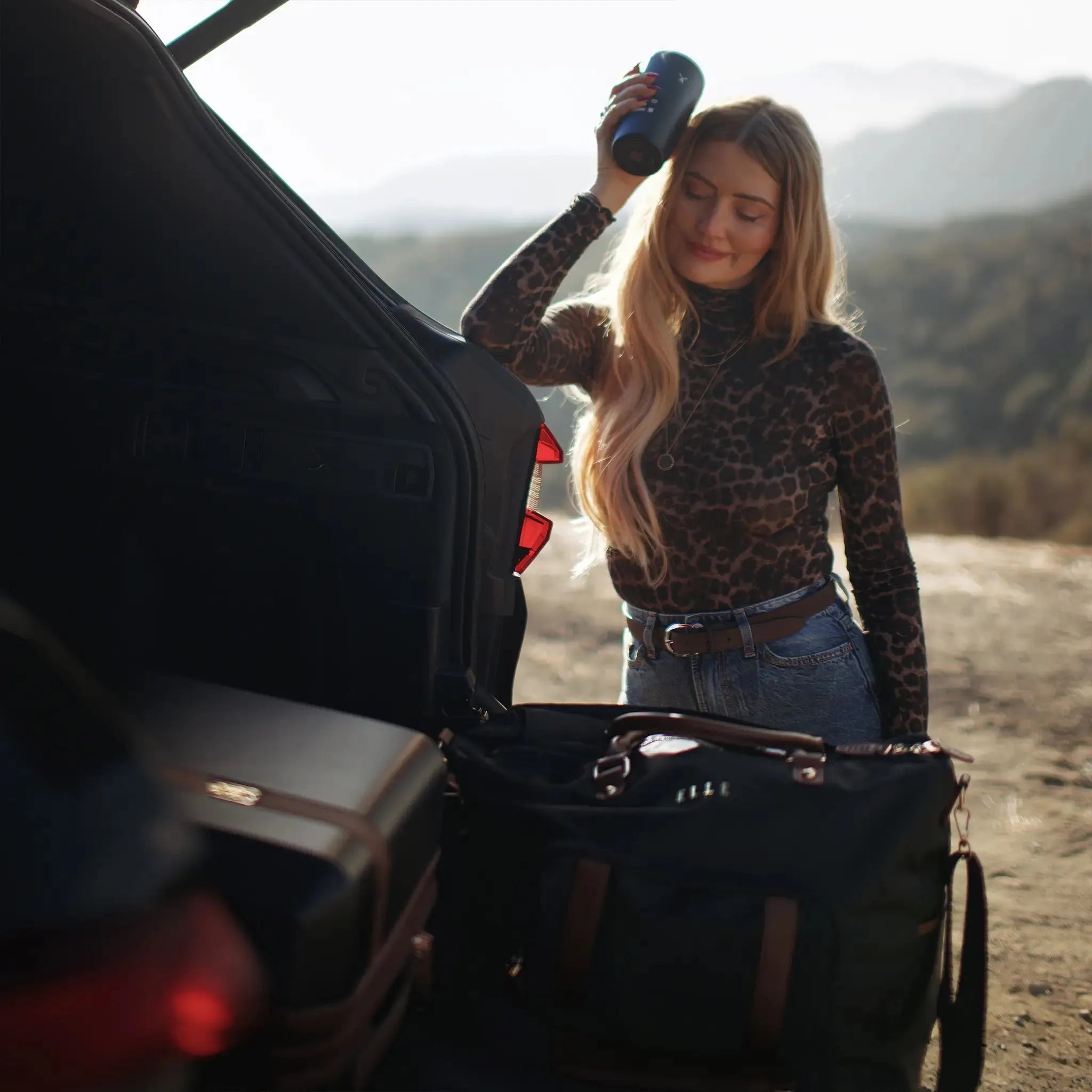 Woman in leopard print top and jeans standing next to an open car trunk with a scenic background.