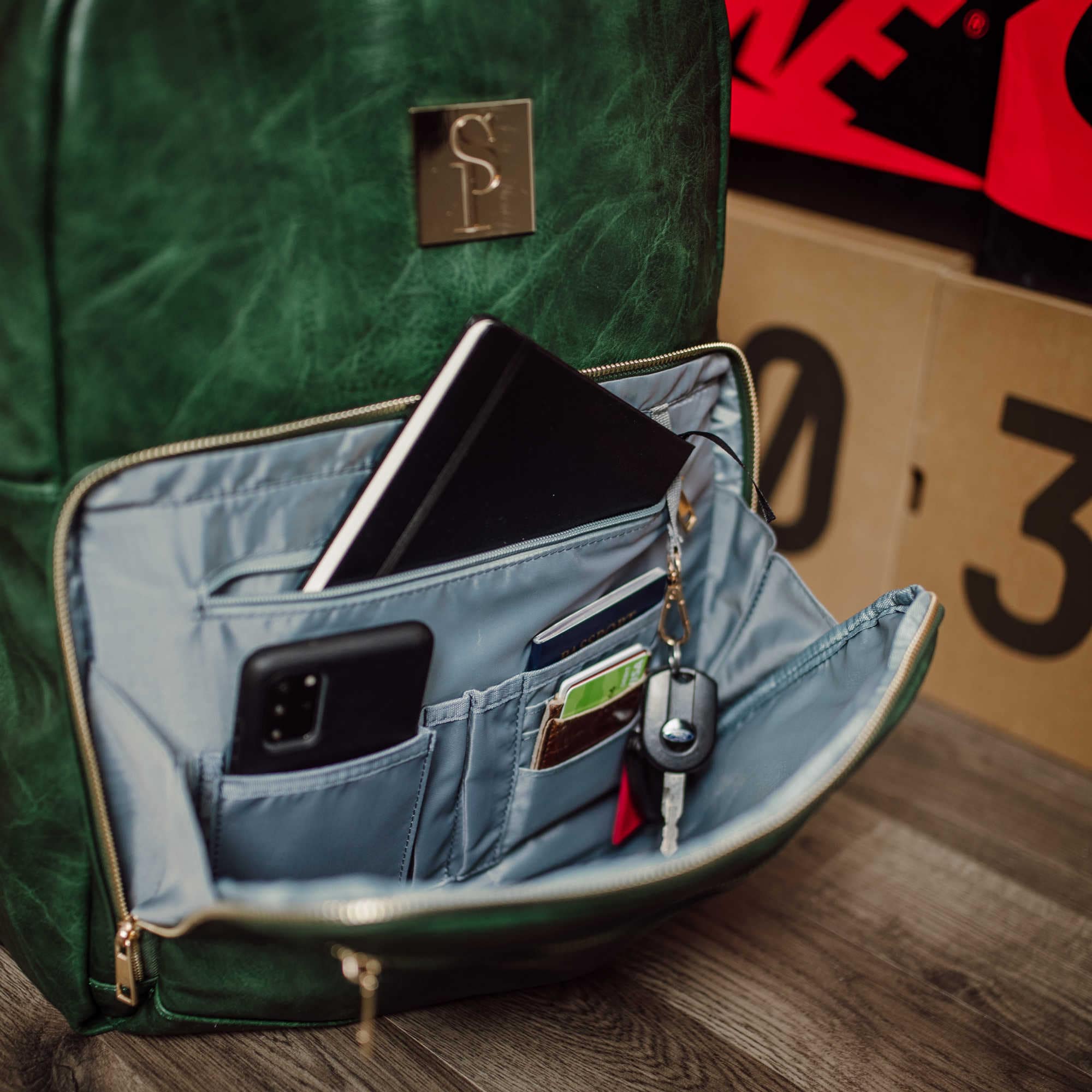 Green backpack with open front pocket showing phone, wallet, and keys on a wooden floor.