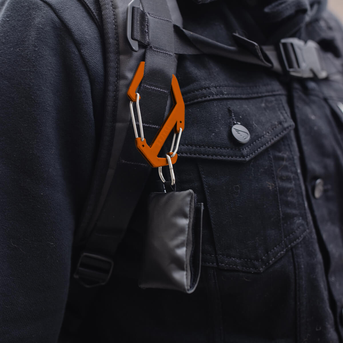 Orange carabiner attached to a black backpack on a person wearing a dark coat.