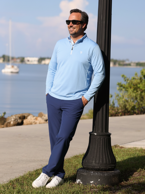 Man in light blue sweater and dark pants leaning against a black lamp post by a waterfront.