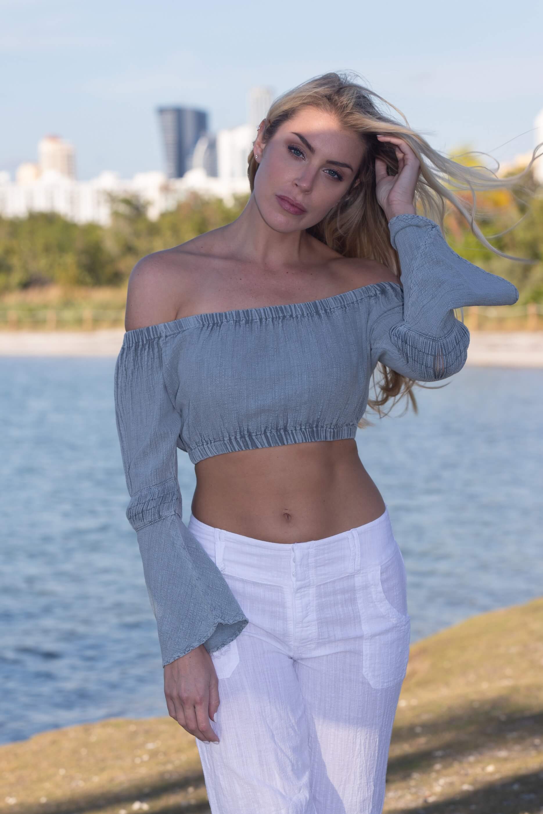 Woman in a blue off-shoulder top and white pants standing by a waterfront with trees and buildings in the background.