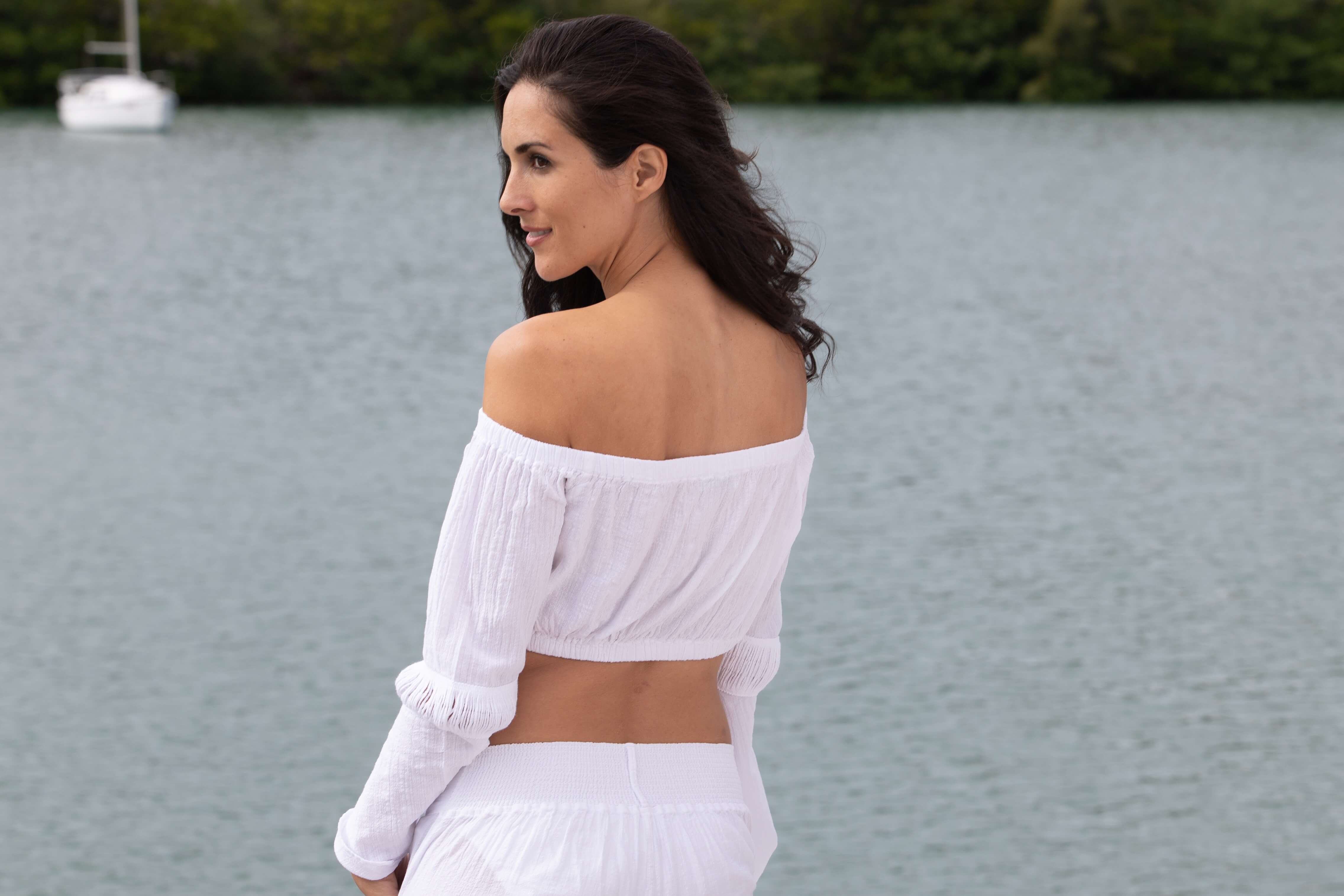 Woman in a white outfit standing by a body of water with a boat in the background.