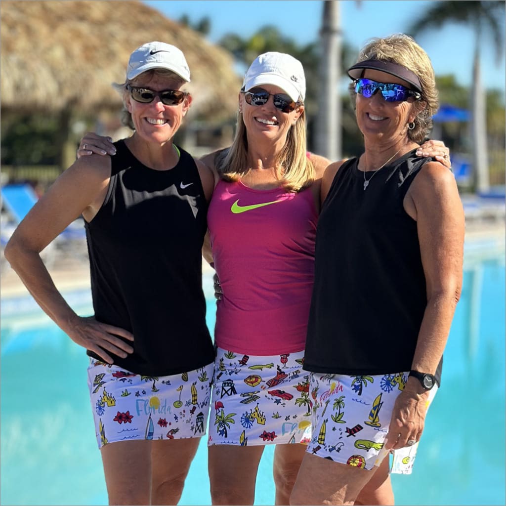 Three women posing together by a pool, wearing sunglasses and colorful shorts.