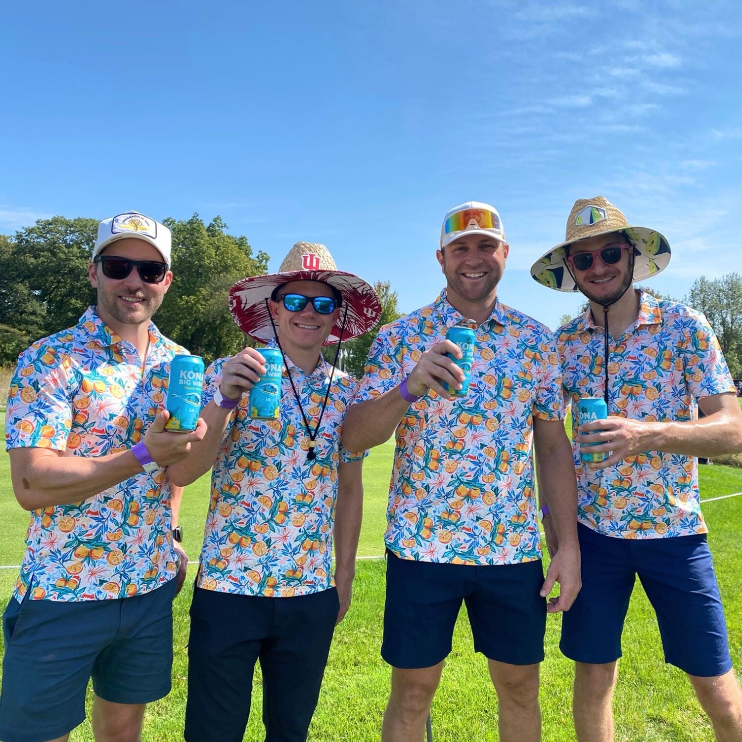 Four men in matching floral shirts and hats holding drinks on a grassy field.