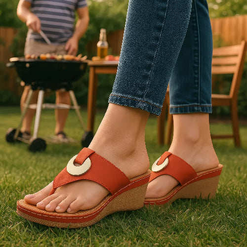 Red sandals with wooden sole worn by a person on grass, with a blurred background of a grill and beer bottle.