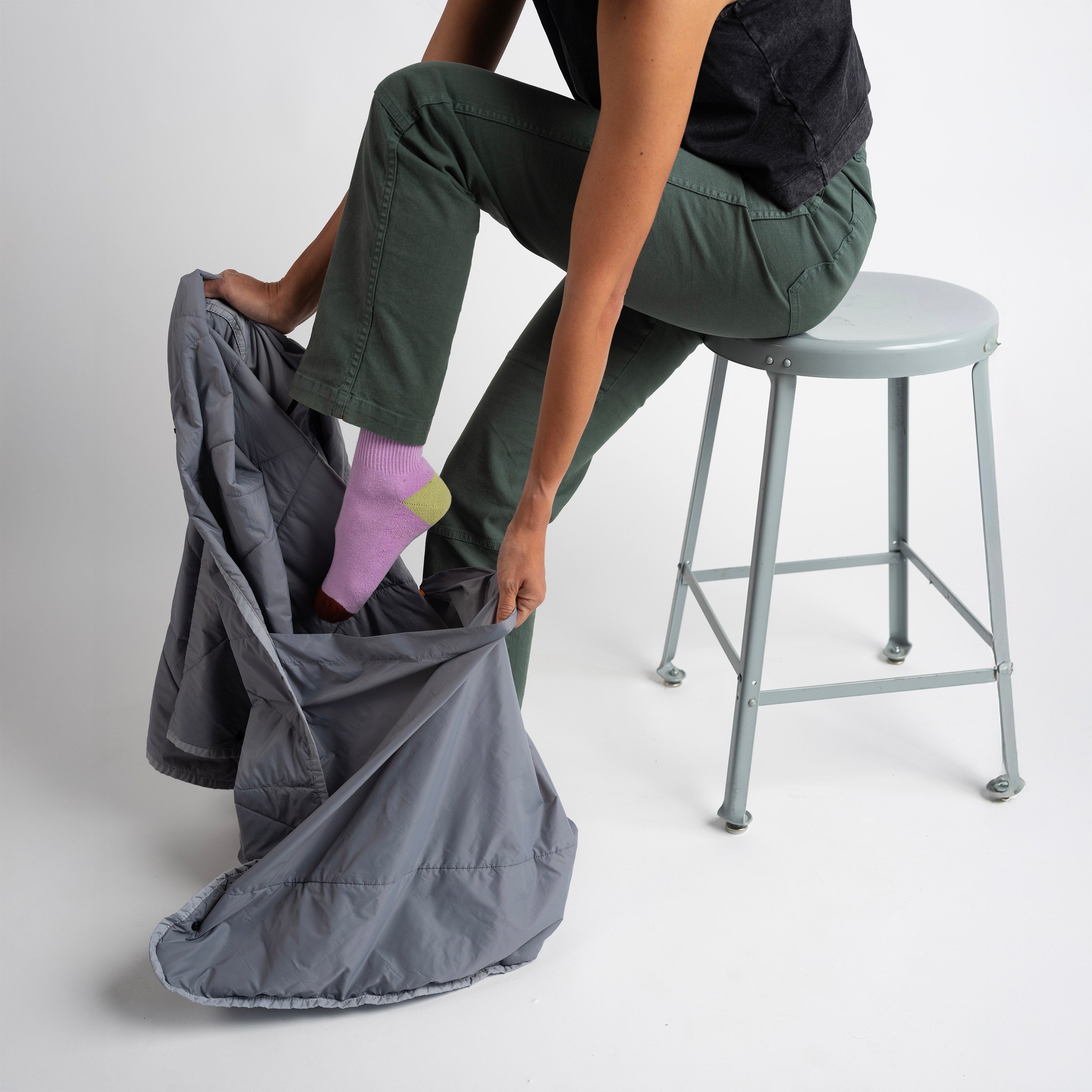 Person sitting on a stool with a gray bag, wearing green pants and pink socks.