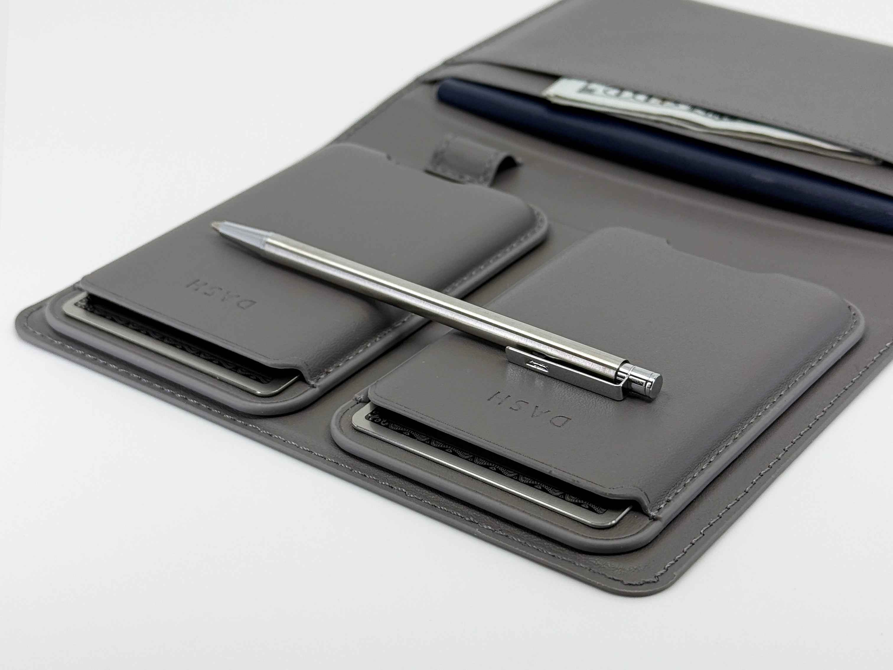 Gray leather case with multiple compartments holding a smartphone, phone case, and two pens on a white background.