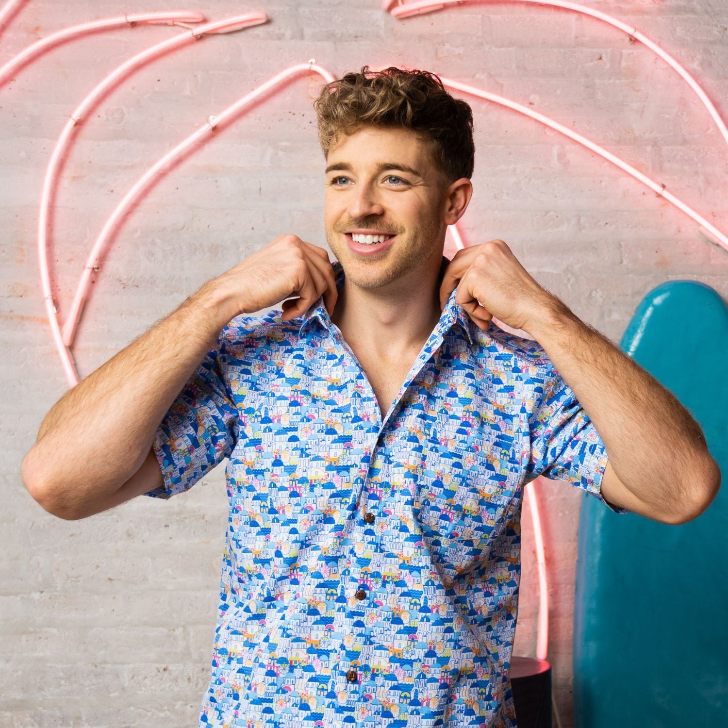 Man adjusting his colorful shirt against a pink neon arch and gray wall.
