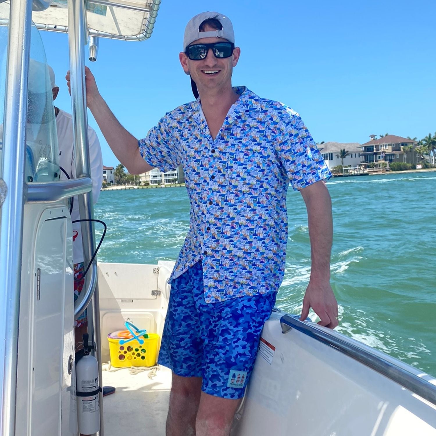 Man on a boat wearing a patterned shirt and blue shorts with a clear blue sky and water background.