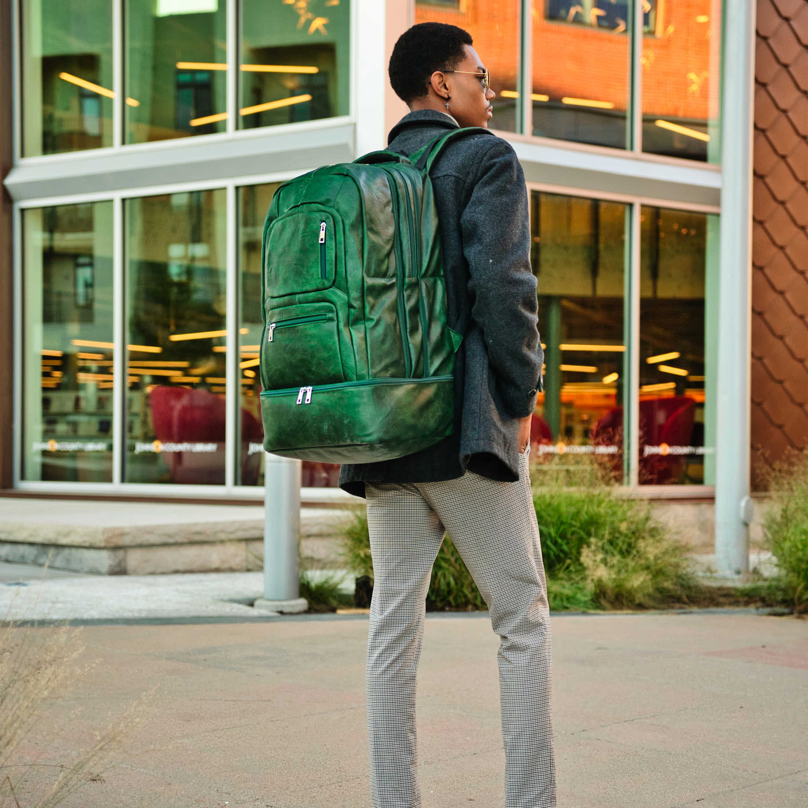 Person wearing a green backpack walking outside a building with large windows.
