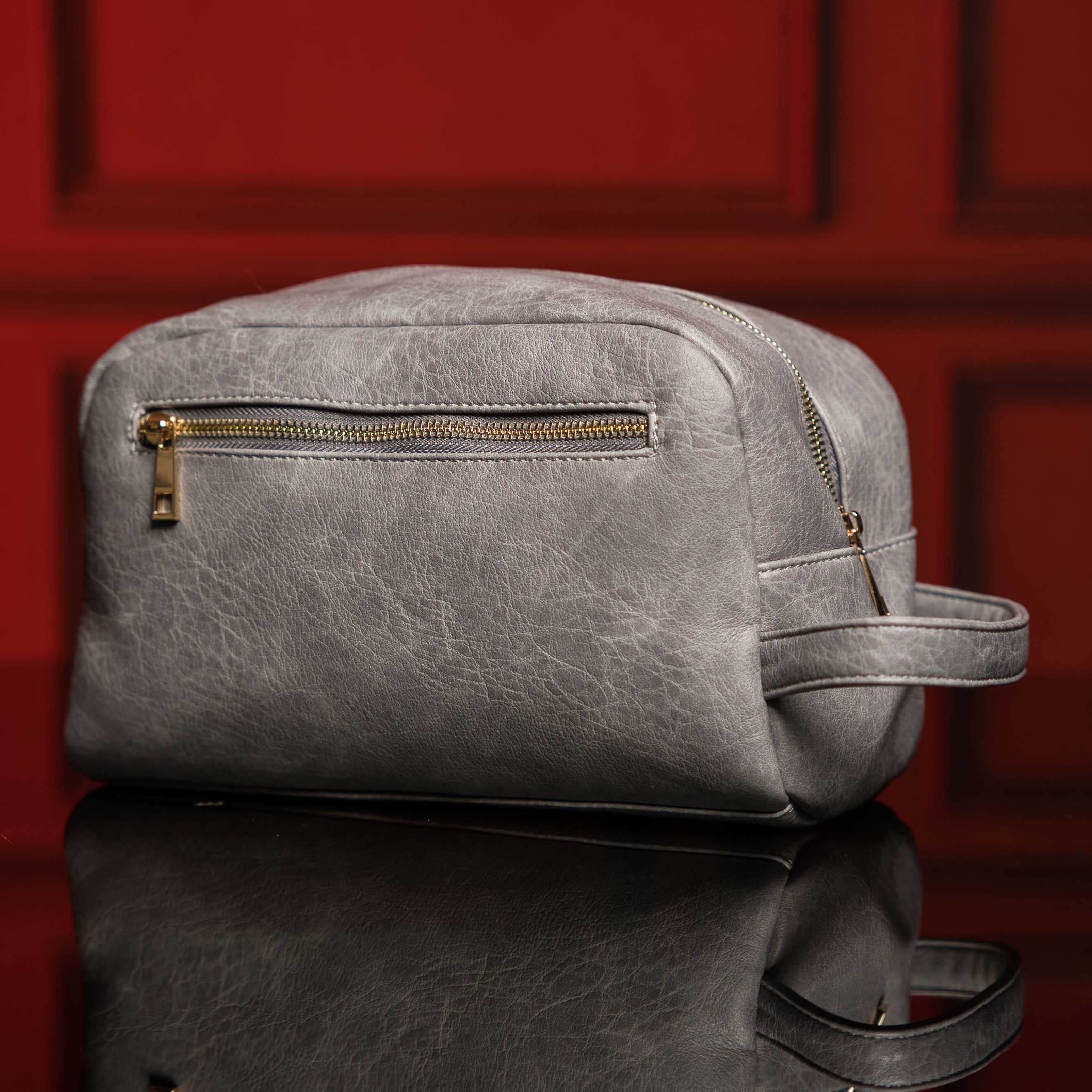 Gray leather dopp kit with gold zipper on a reflective surface with a red background