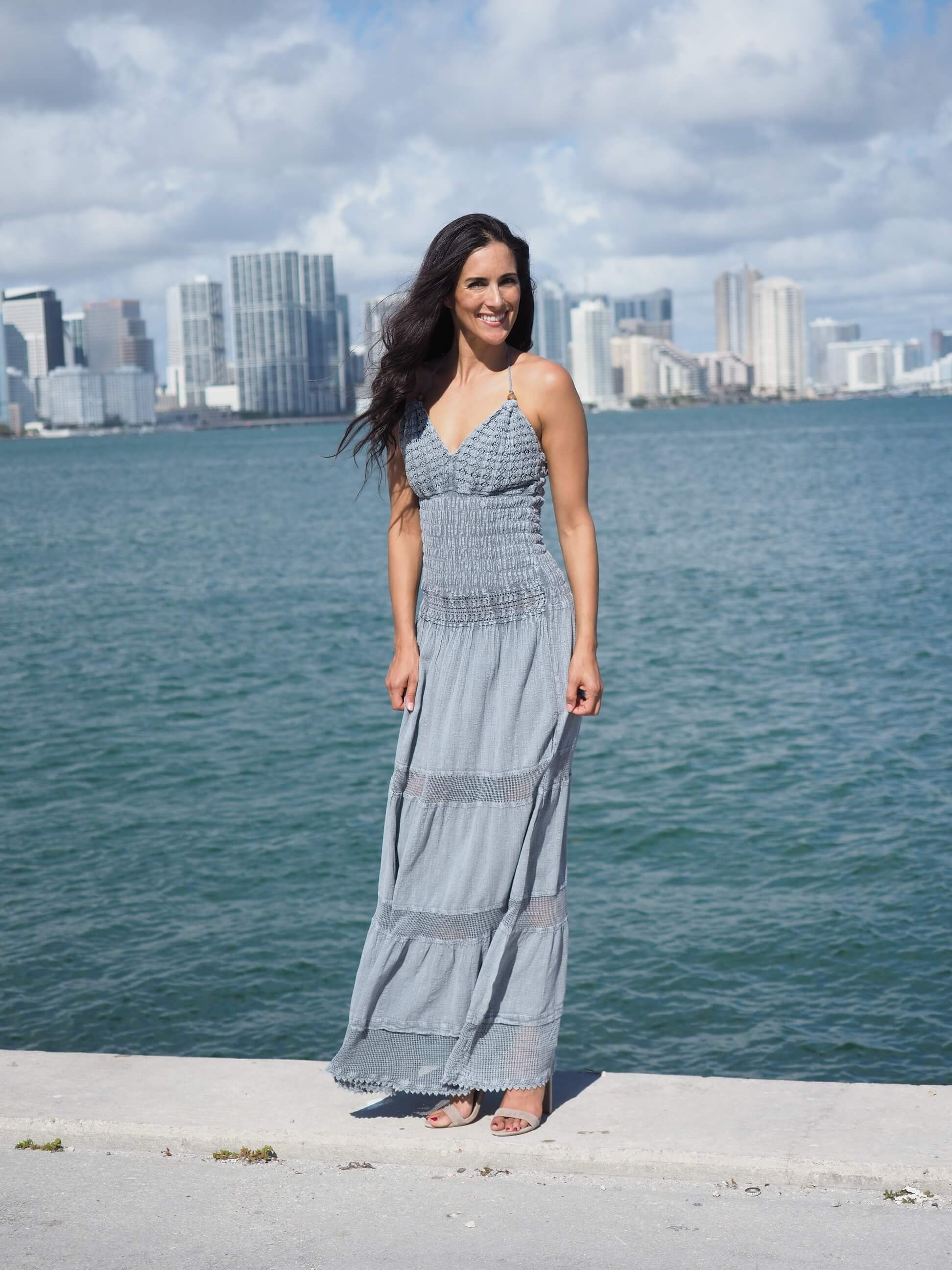 Woman in a gray dress standing by a waterfront with a city skyline in the background