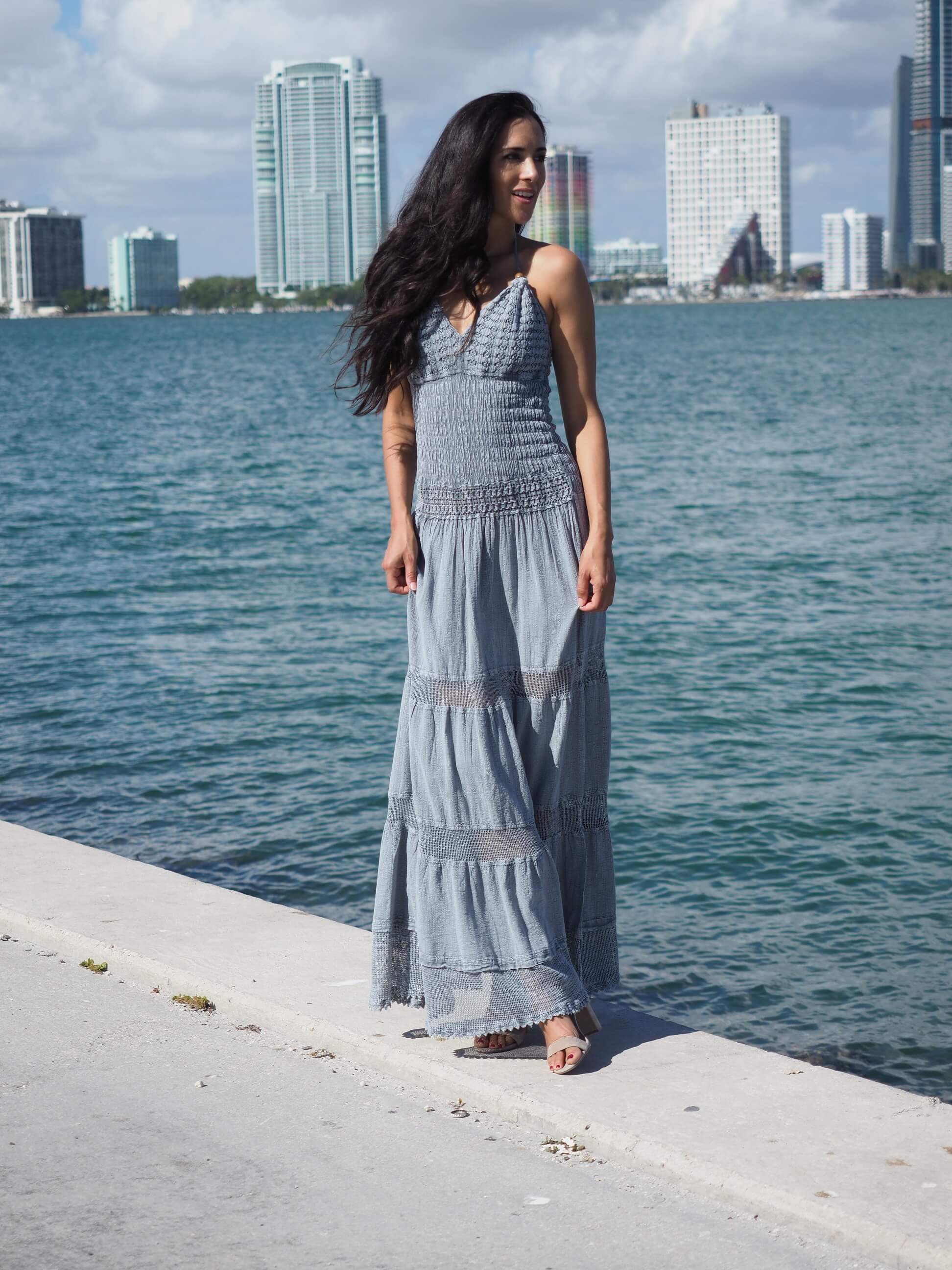Woman in a long gray dress standing by a waterfront with city skyline in the background