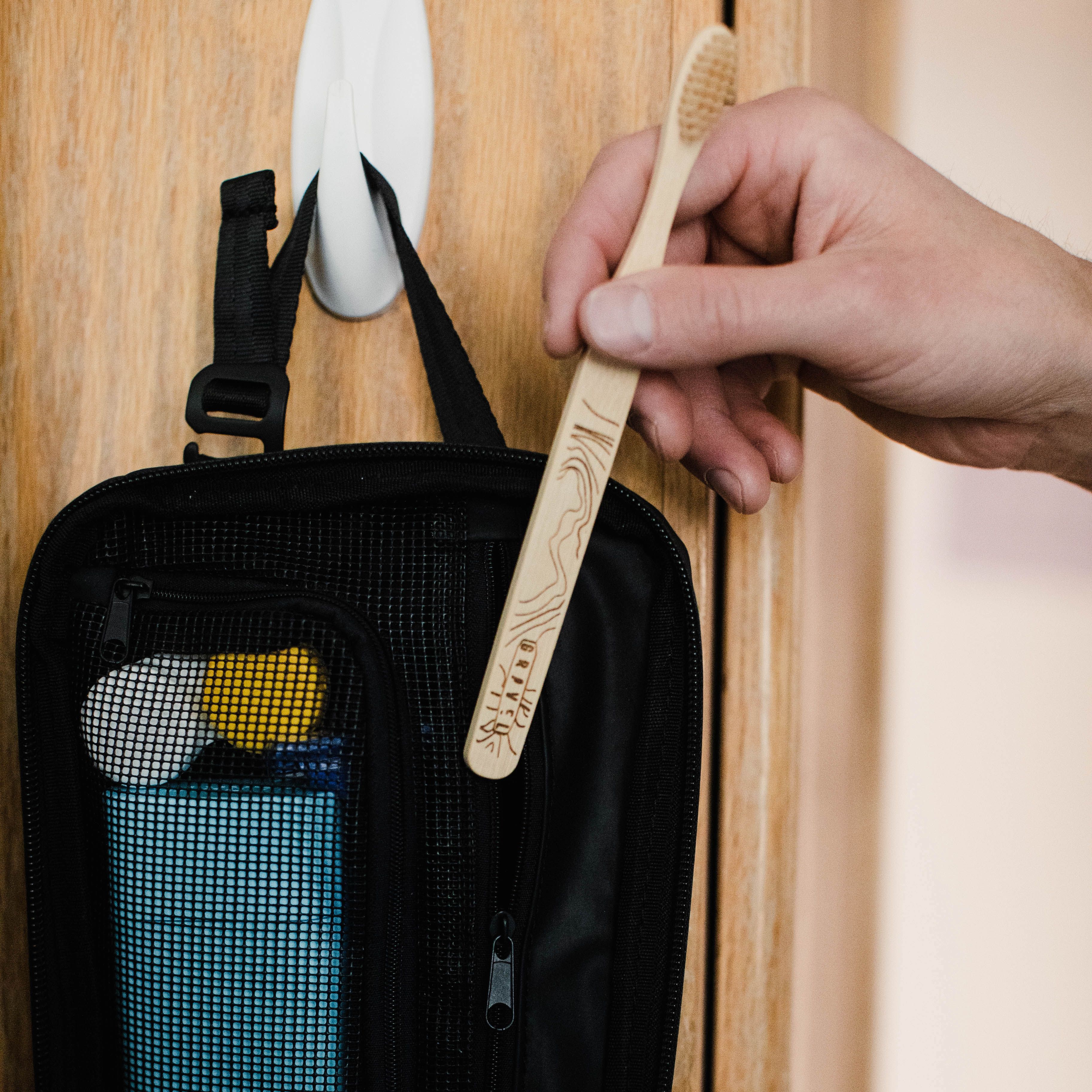 Person holding a wooden toothbrush next to a black toiletry bag with travel items on a wooden surface.