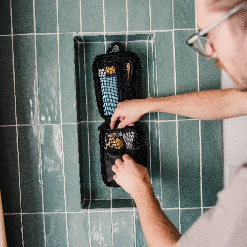 Person organizing toiletries in a black travel pouch against a green tiled wall.