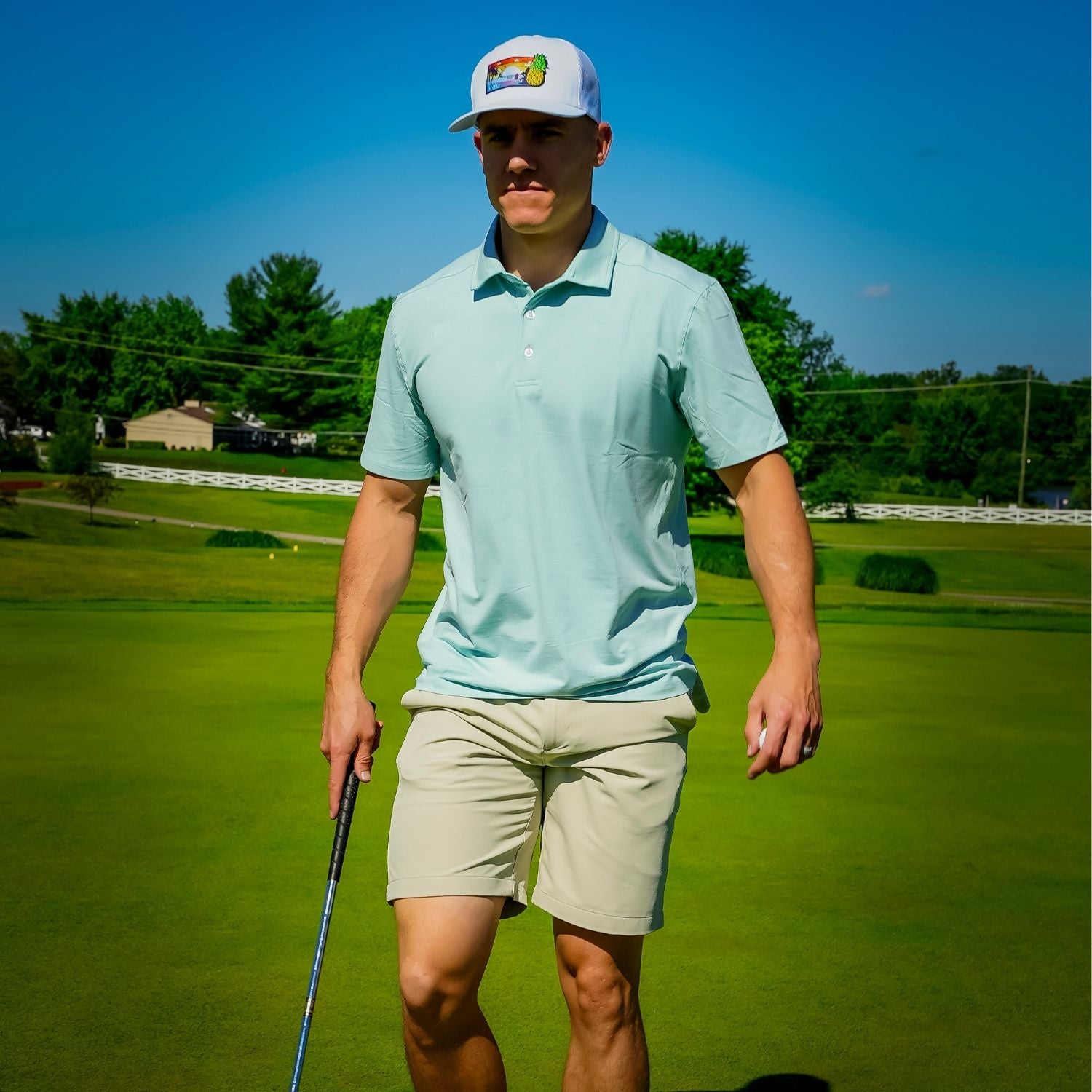 Man on a golf course wearing a light blue shirt and beige shorts, holding a golf club.