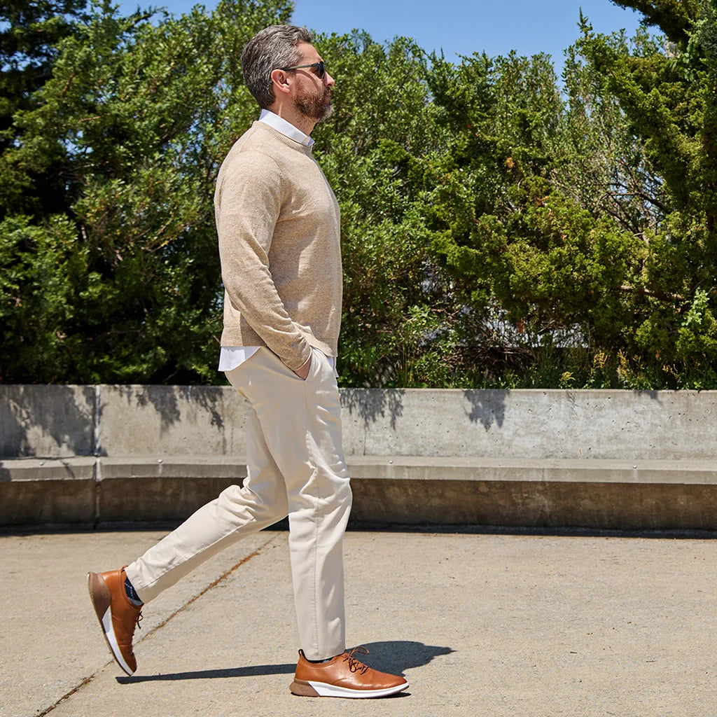 Man walking outdoors wearing a beige sweater, white pants, and brown shoes.