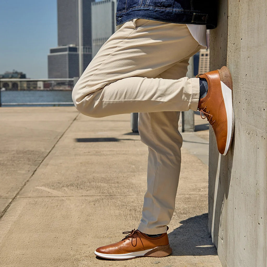 Person wearing tan pants and brown leather shoes leaning against a concrete wall with a cityscape in the background.