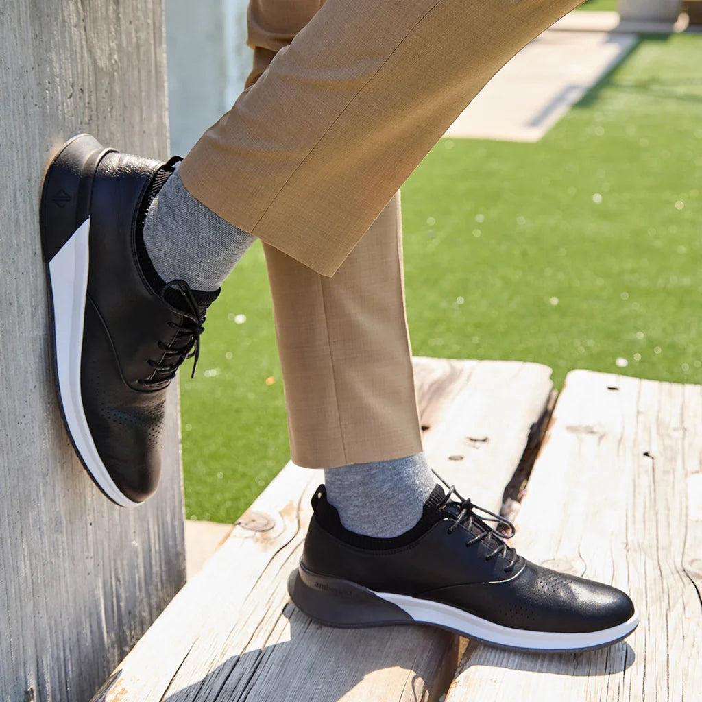 Person wearing black leather shoes with white soles, standing on a wooden bench.