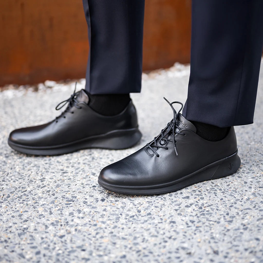 Black leather shoes worn with dark pants on a textured pavement.