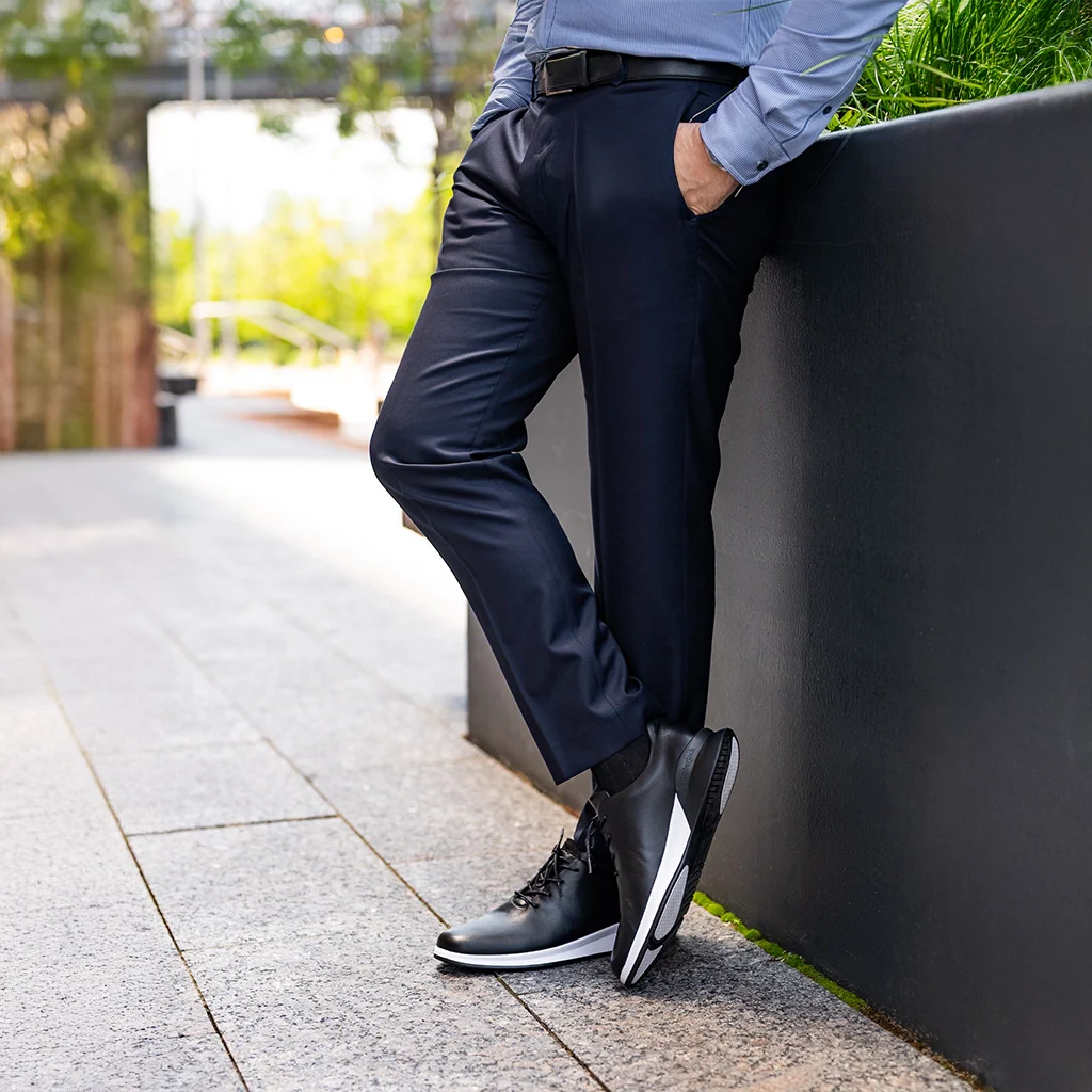 Person wearing navy pants and black shoes leaning against a wall outdoors.