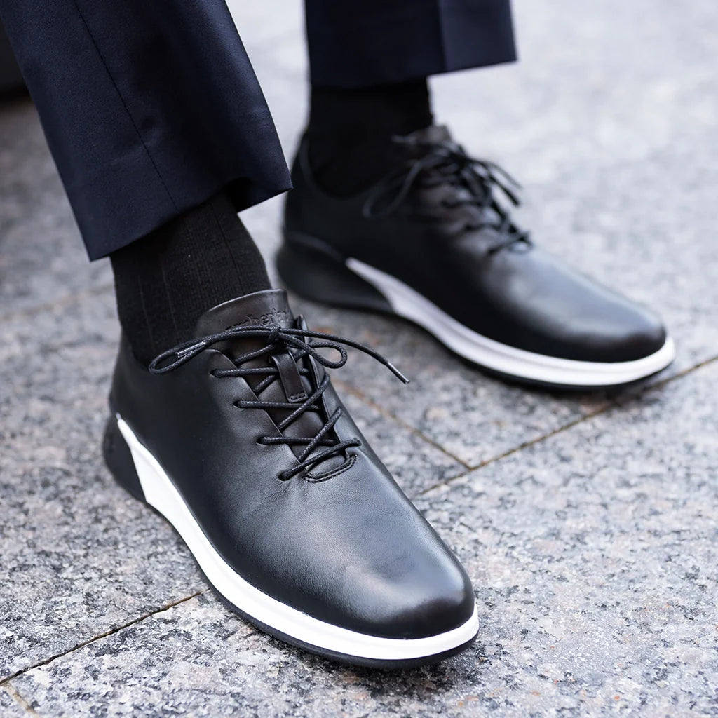 Black leather shoes with white soles worn by a person on a pavement.