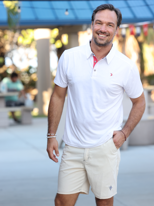Man wearing a white polo shirt and beige shorts standing outdoors.