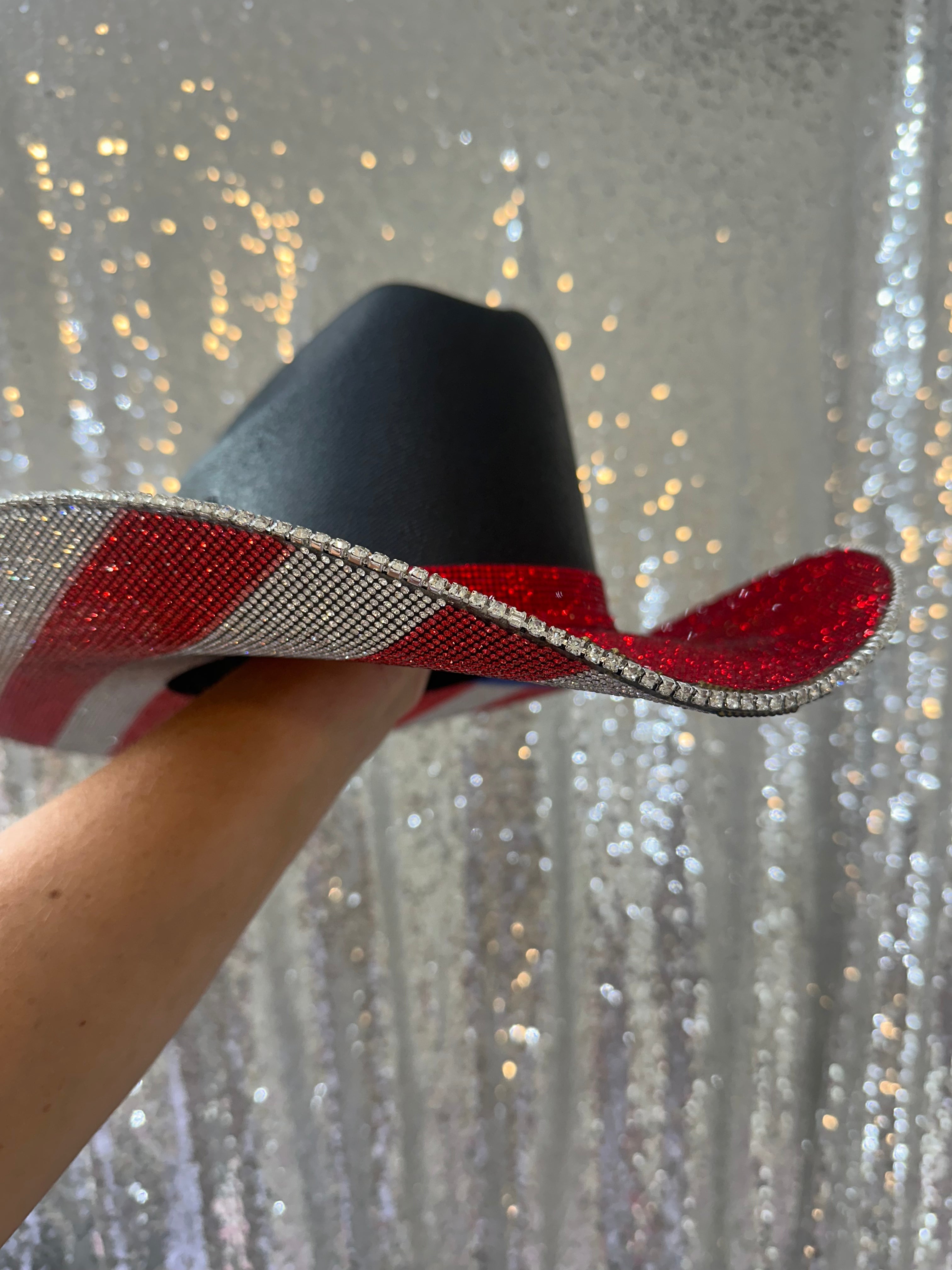 Black cowboy hat with red band held by a hand against a glittery silver background