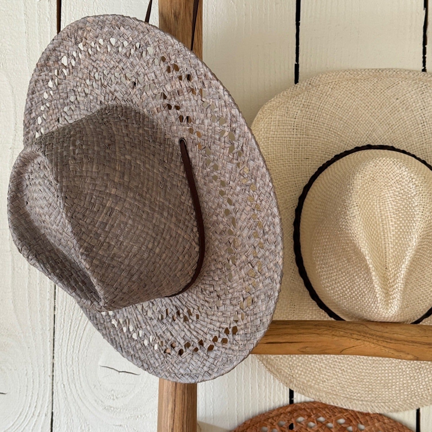 Two straw hats, one brown and one beige, hanging on a wooden rack against a white wooden wall.