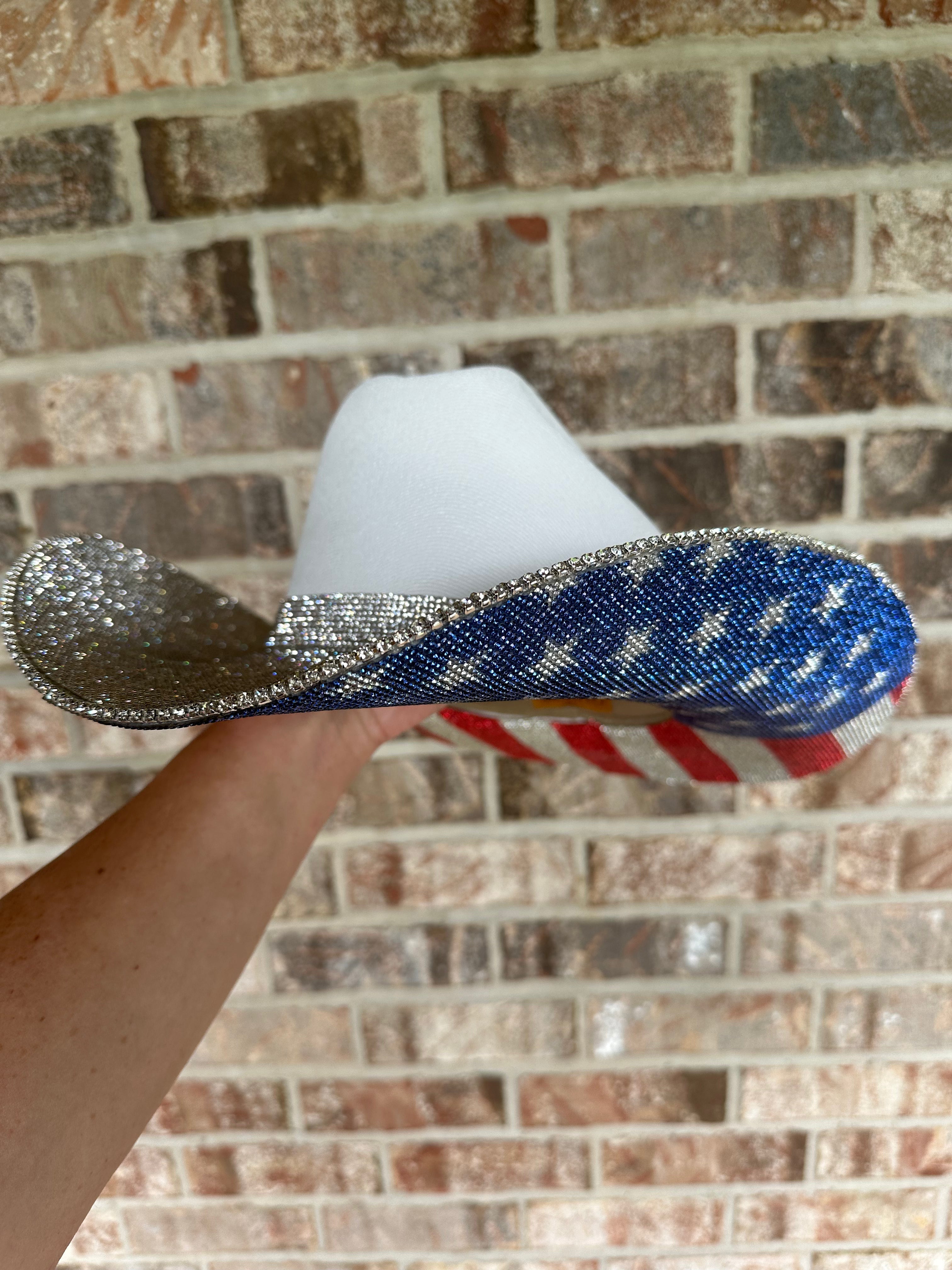 Hand holding a decorative hat with a white center, blue and red brim, against a brick wall.