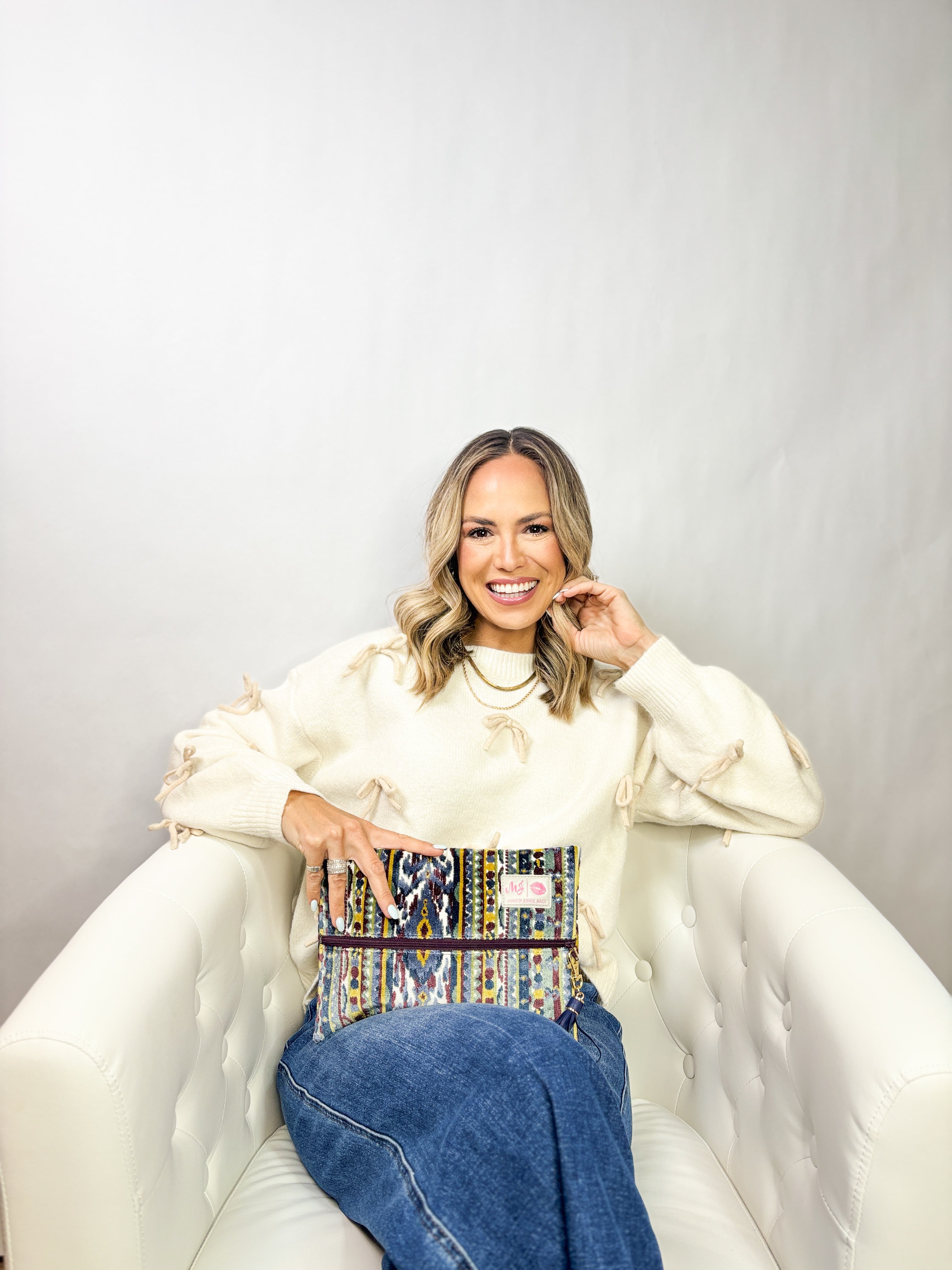Woman sitting on a white couch holding a colorful handbag against a plain background