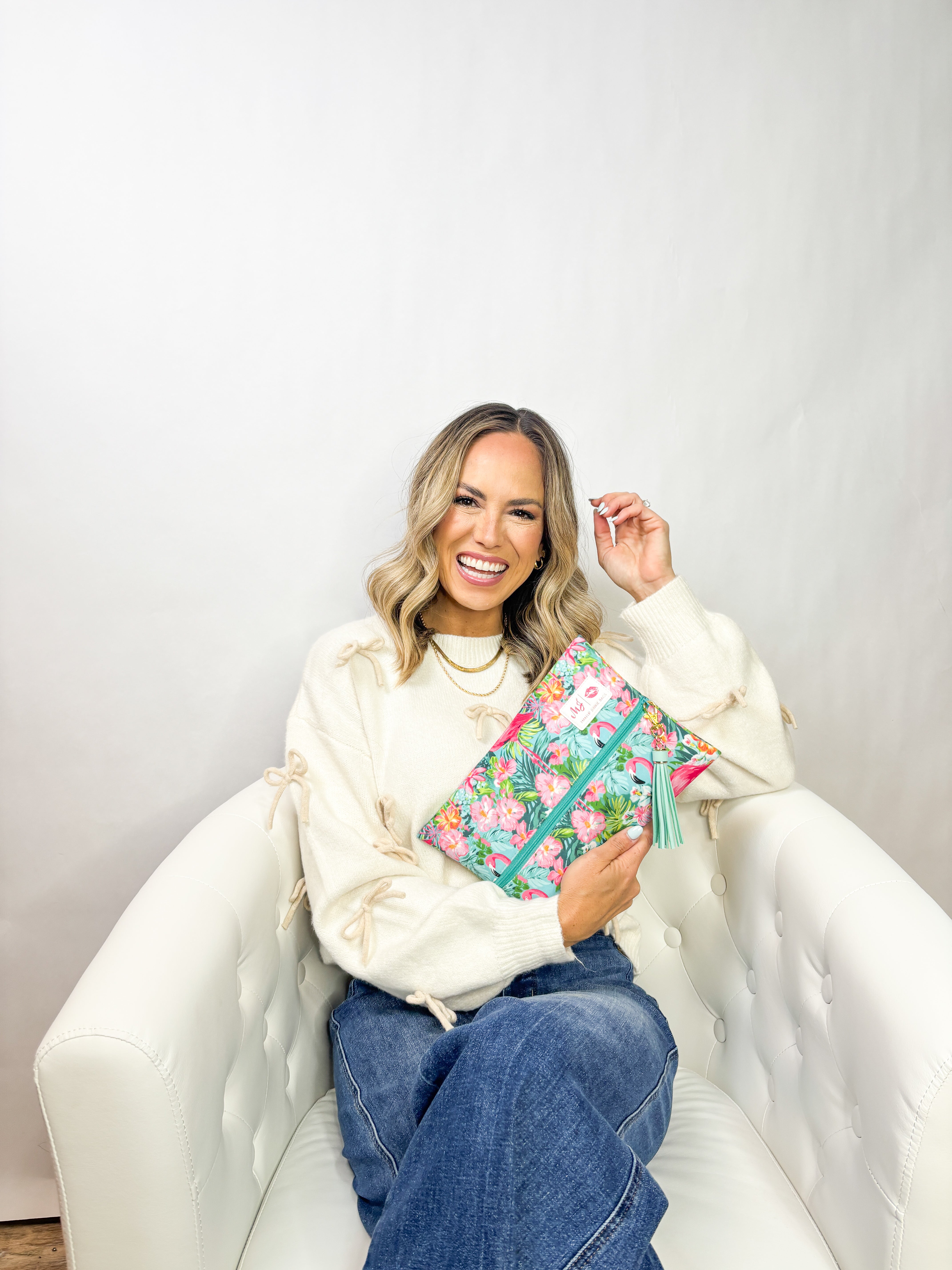 Woman sitting on a white chair holding a colorful floral clutch against a plain background