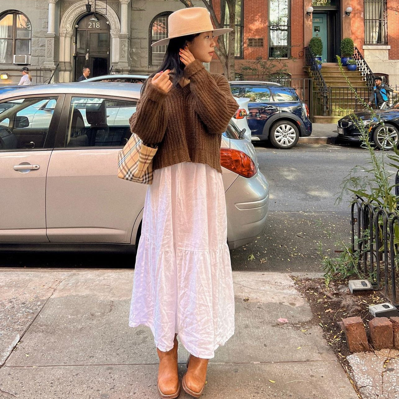 Woman in a brown sweater and white dress standing on a city street.