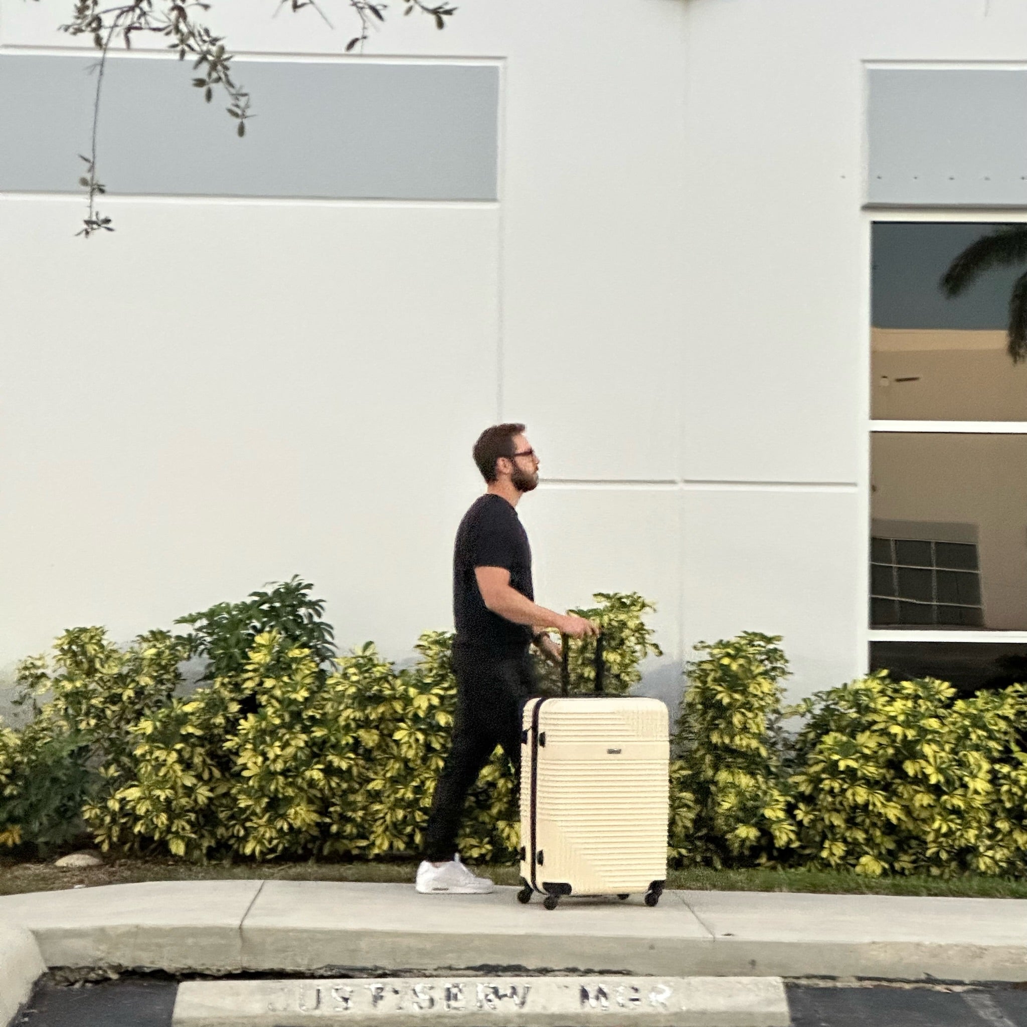 Man pulling a beige suitcase on a sidewalk with greenery and a building in the background