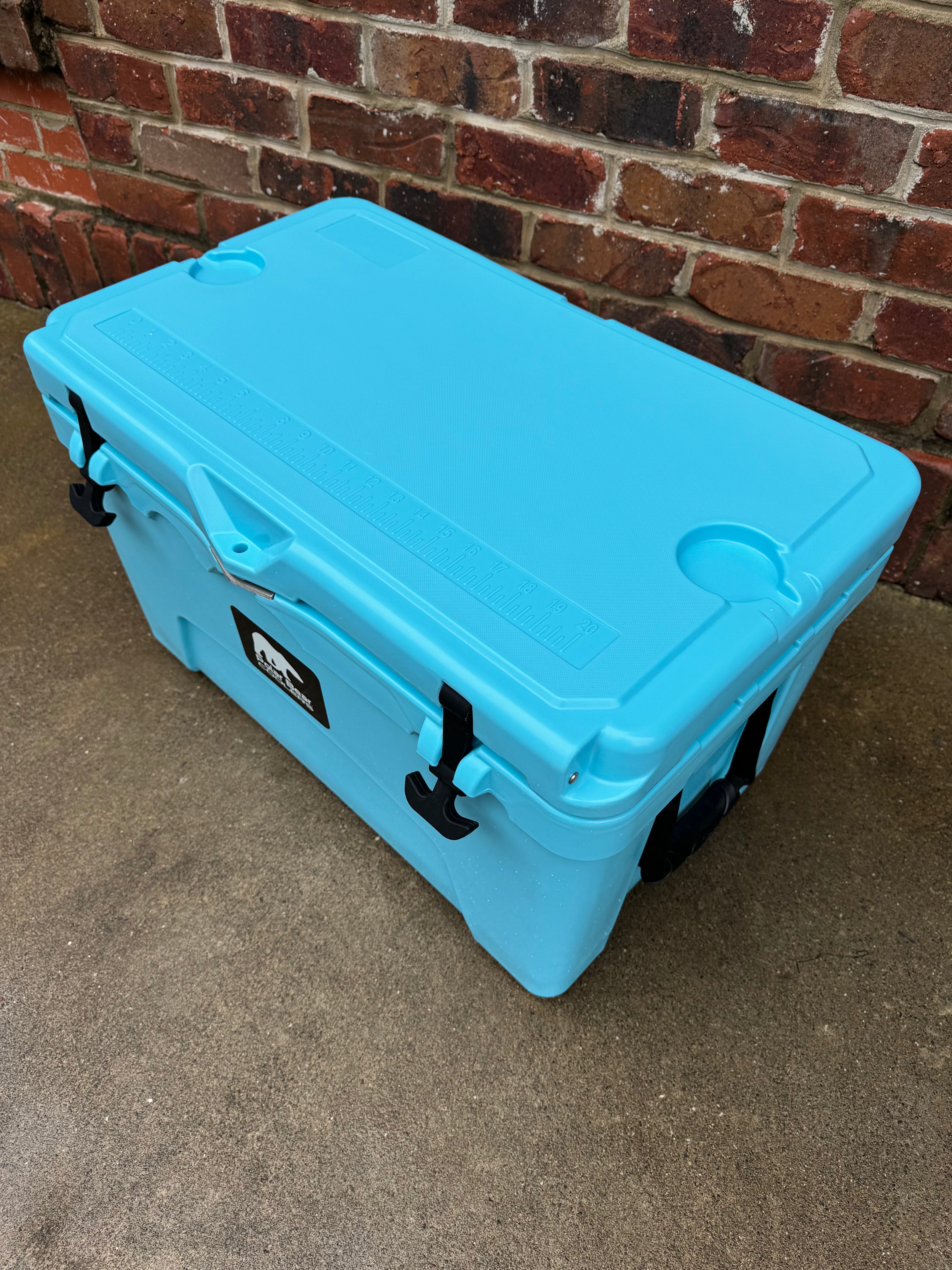 Blue cooler box with a logo on a concrete floor against a brick wall.