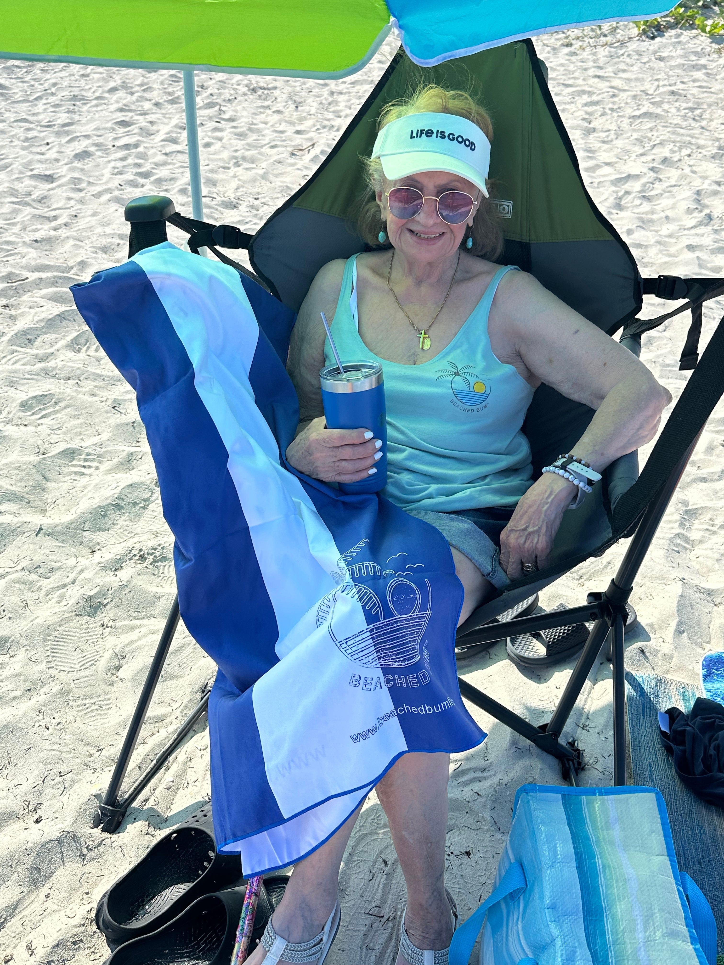 Woman sitting on a beach chair with a towel and drink, wearing a 'Life is Good' headband.
