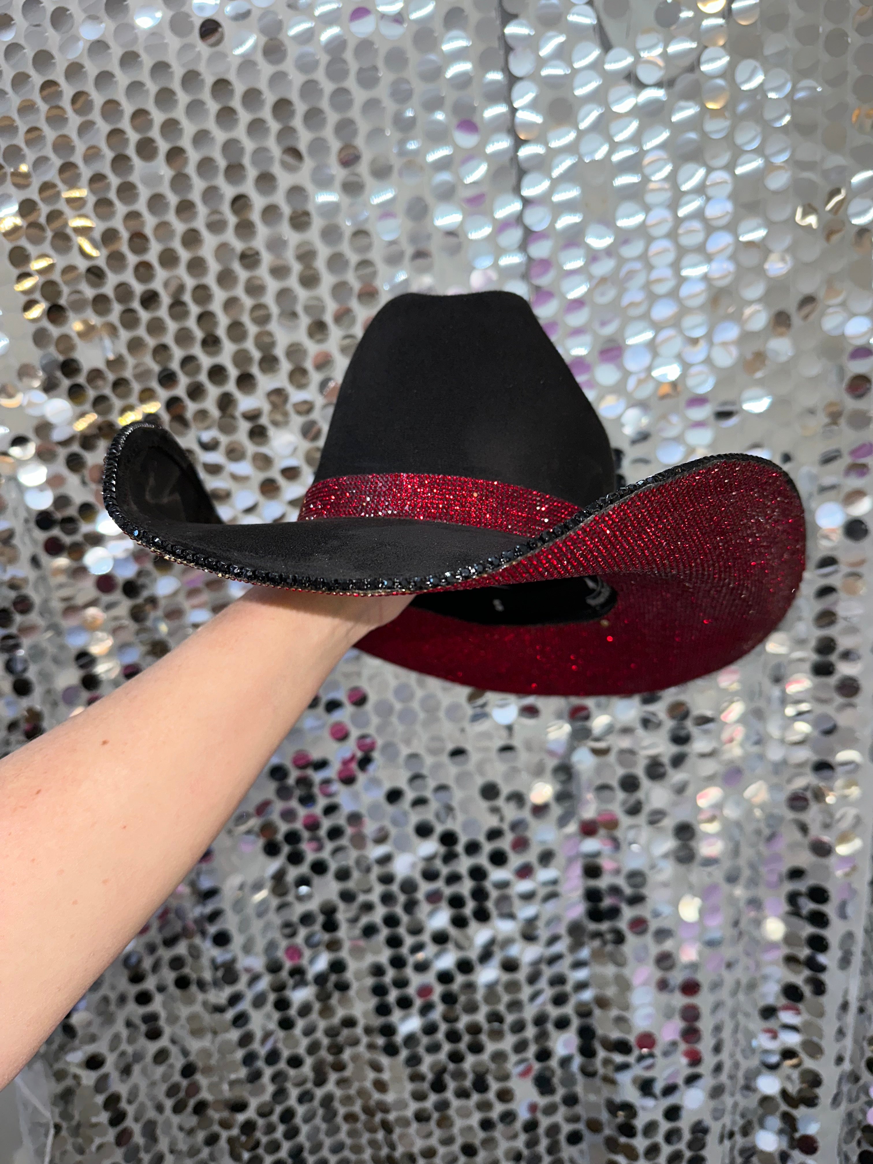 Black and red glittery cowboy hat held by a hand against a silver sequin background