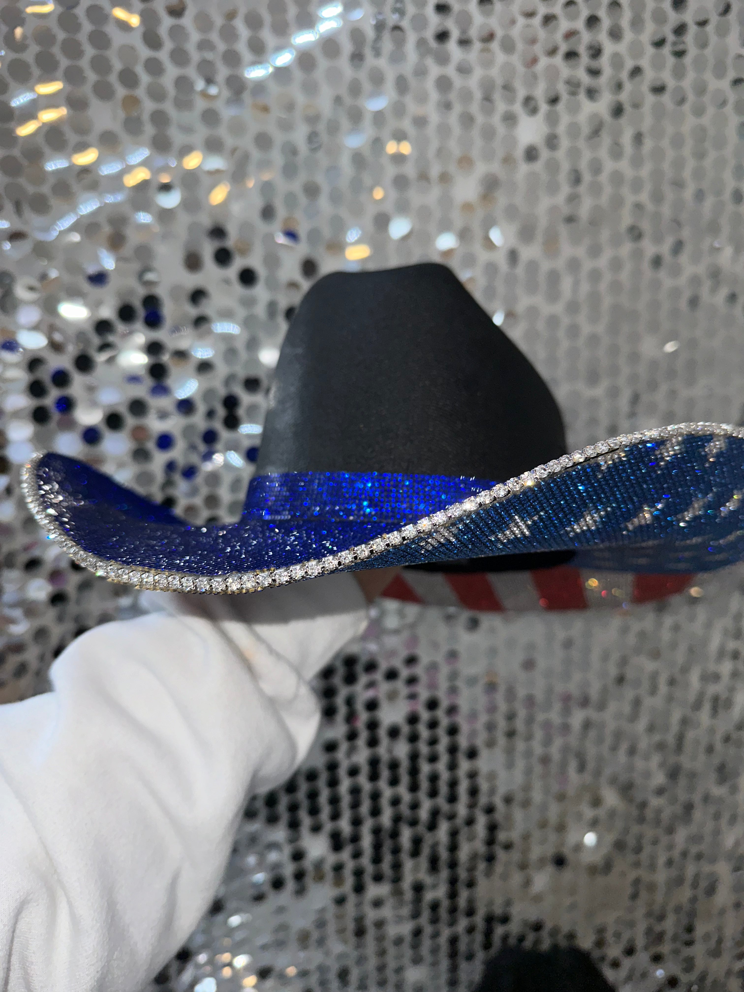 Black cowboy hat with blue brim held by a gloved hand against a glittery background