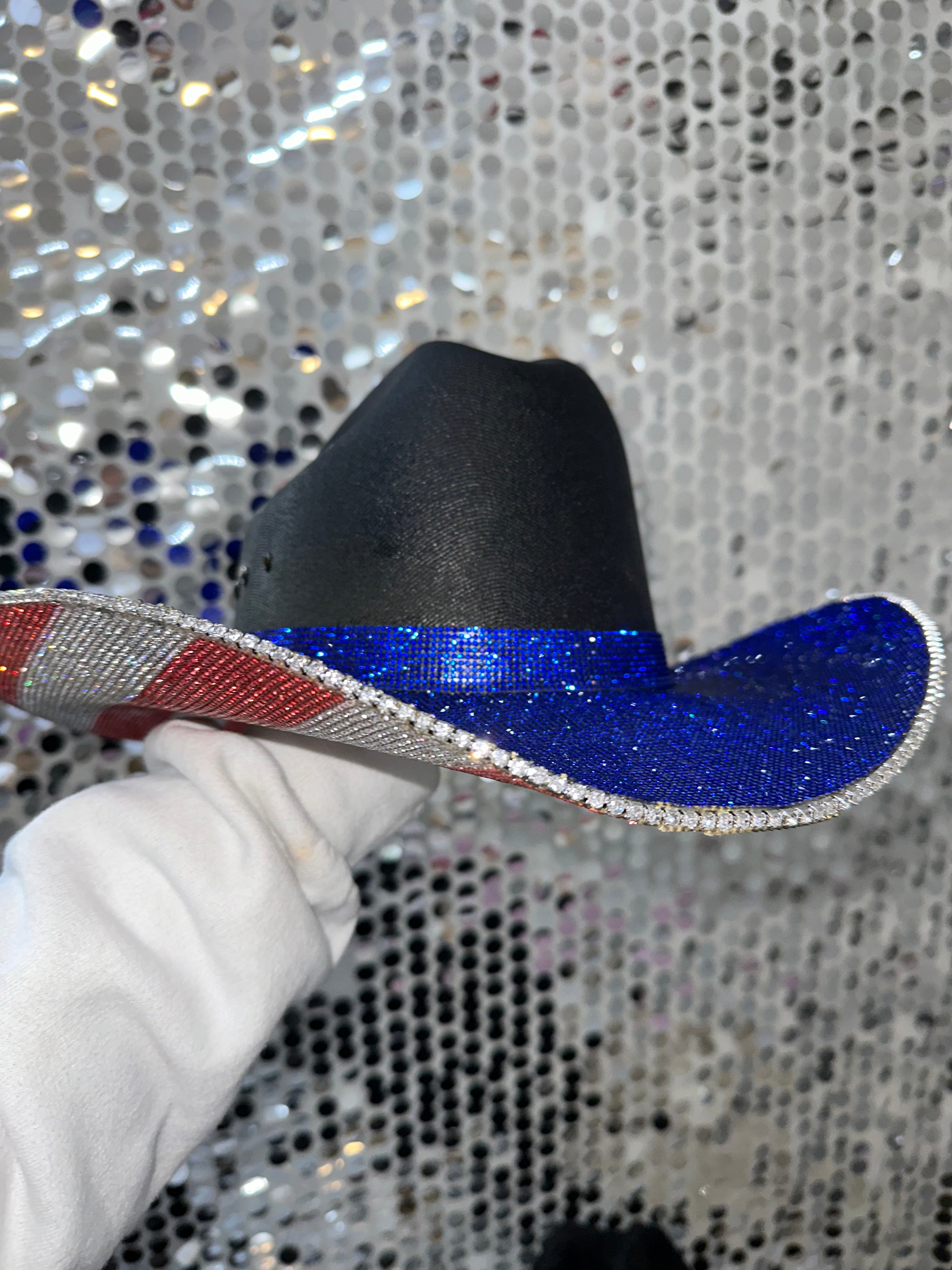 Black cowboy hat with a blue brim held by a gloved hand against a sequined background