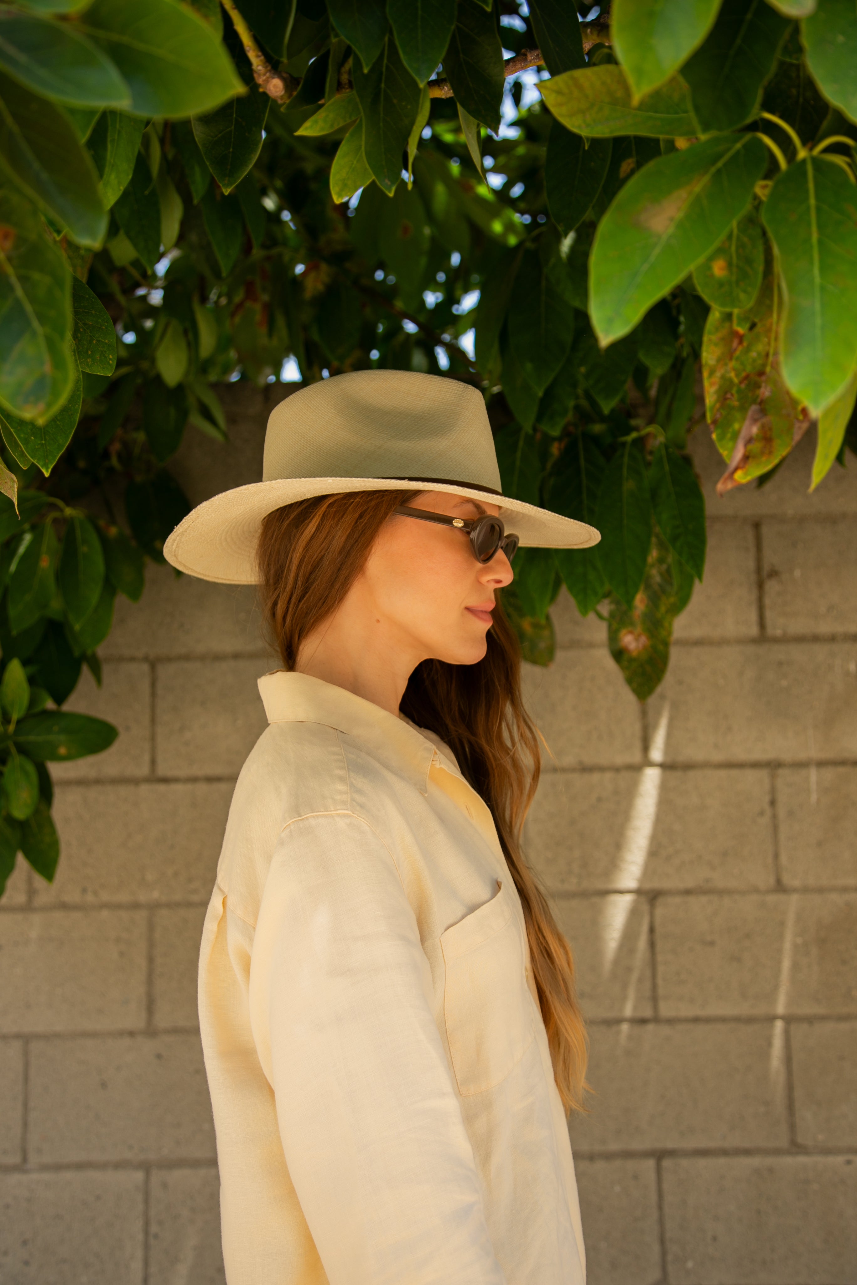 Woman wearing a beige hat and sunglasses, standing against a brick wall with green foliage.