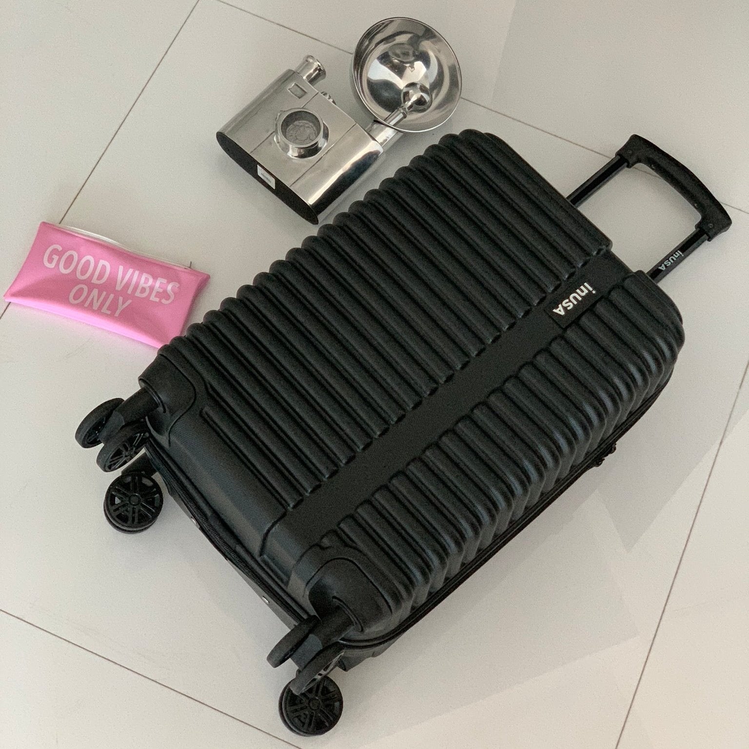 Black suitcase with a camera, pink pouch, and silver object on a light gray floor.